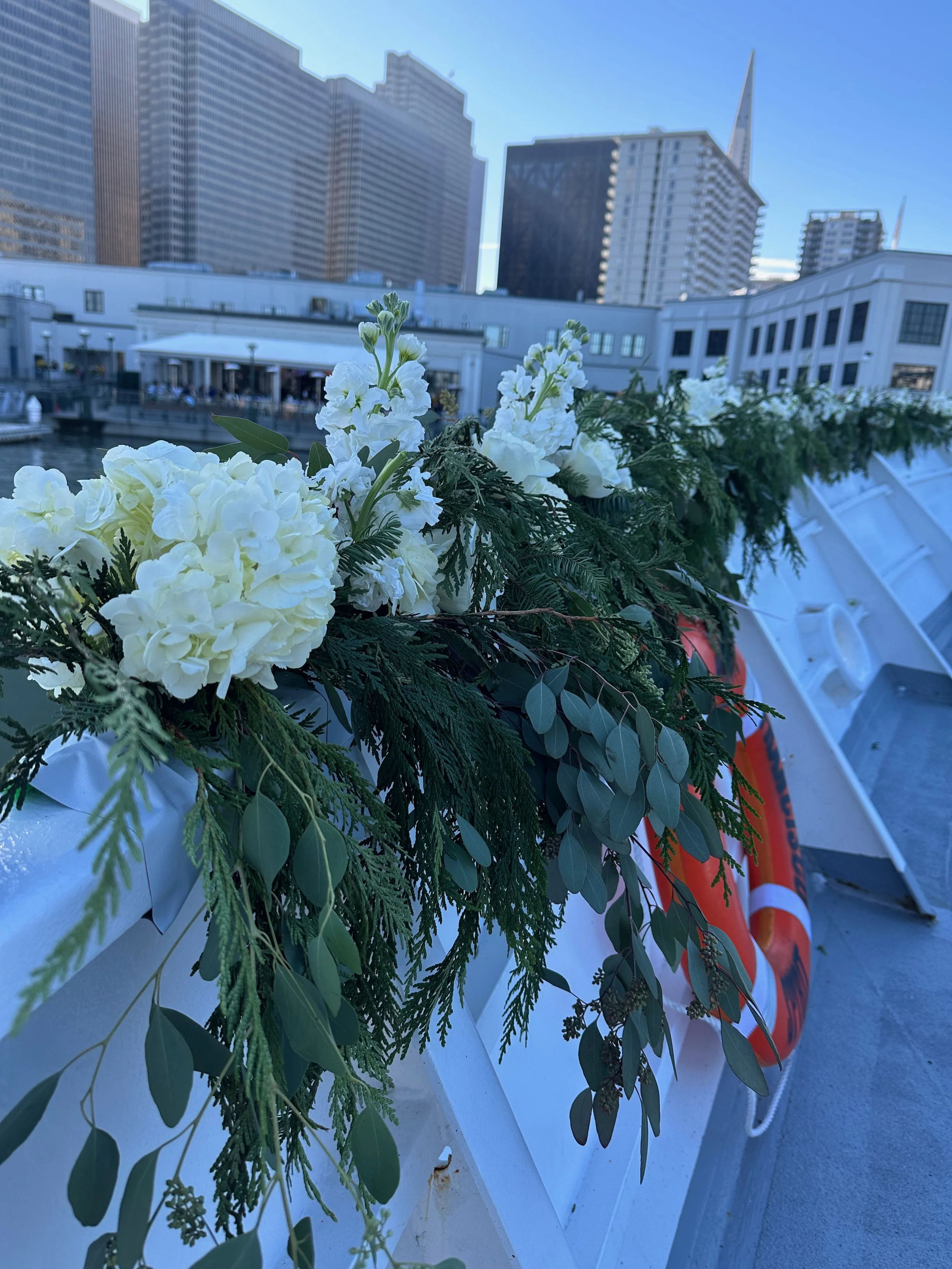 Close-up of white flowers and green foliage on a boat railing with city buildings and cranes in the background.