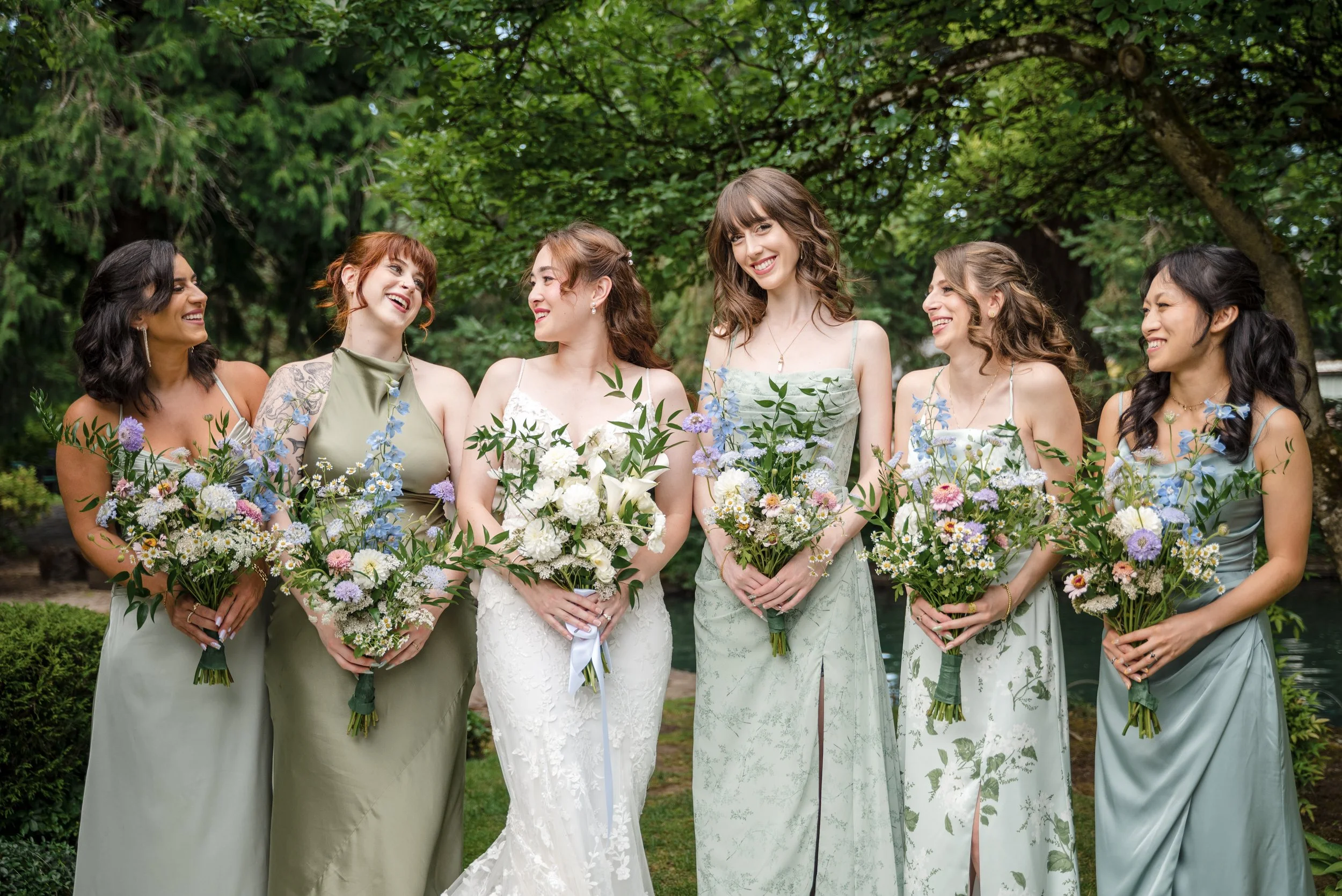 Group of six women dressed in formal attire, holding bouquets of flowers, standing outdoors in a lush green garden.