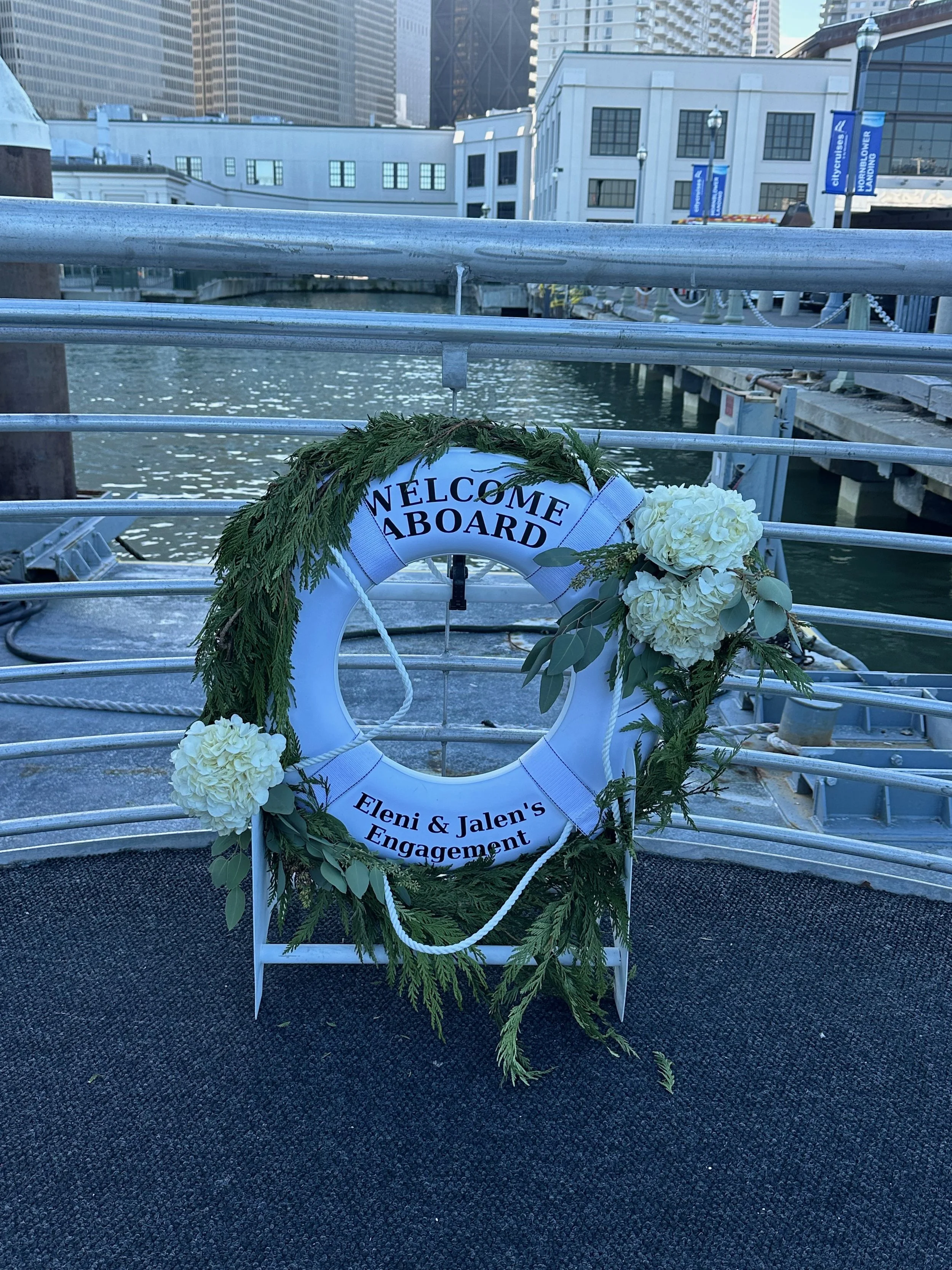 A decorated life preserver on a boat dock reads 'Welcome Aboard' and 'Eleni & Jalen's Engagement,' adorned with greenery and white flowers.