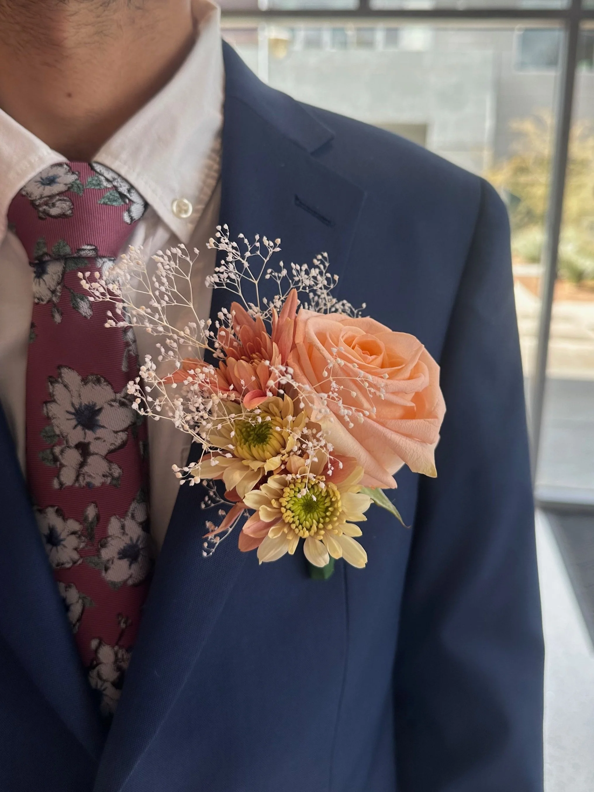 A man in a navy suit with a white shirt and floral tie, wearing a pink rose boutonniere with additional small flowers and baby's breath.