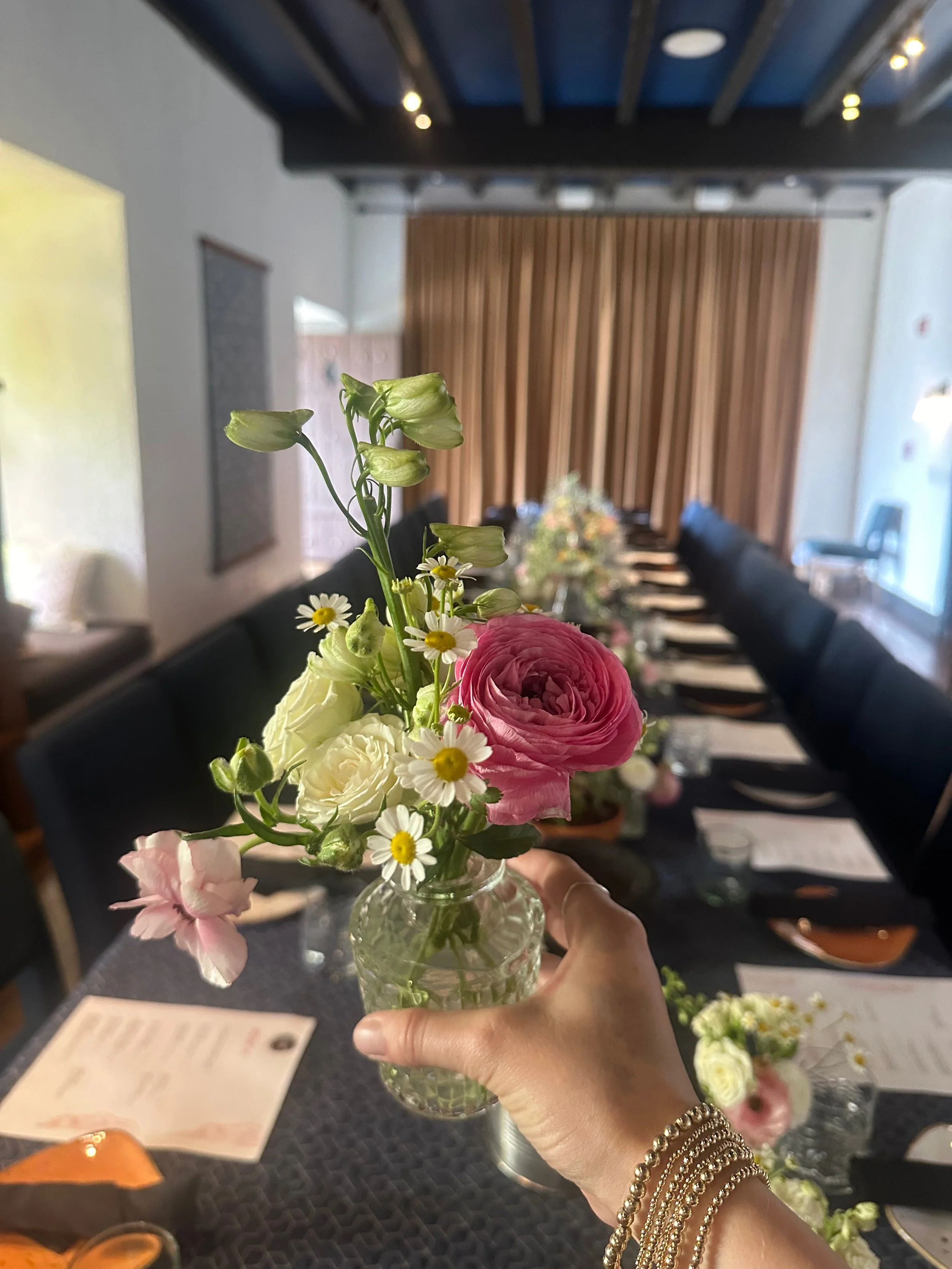 Person holding a small glass vase with pink and white flowers and greenery, over a decorated dining table with place cards, silverware, and other floral arrangements in a room with wood paneling and black chairs.