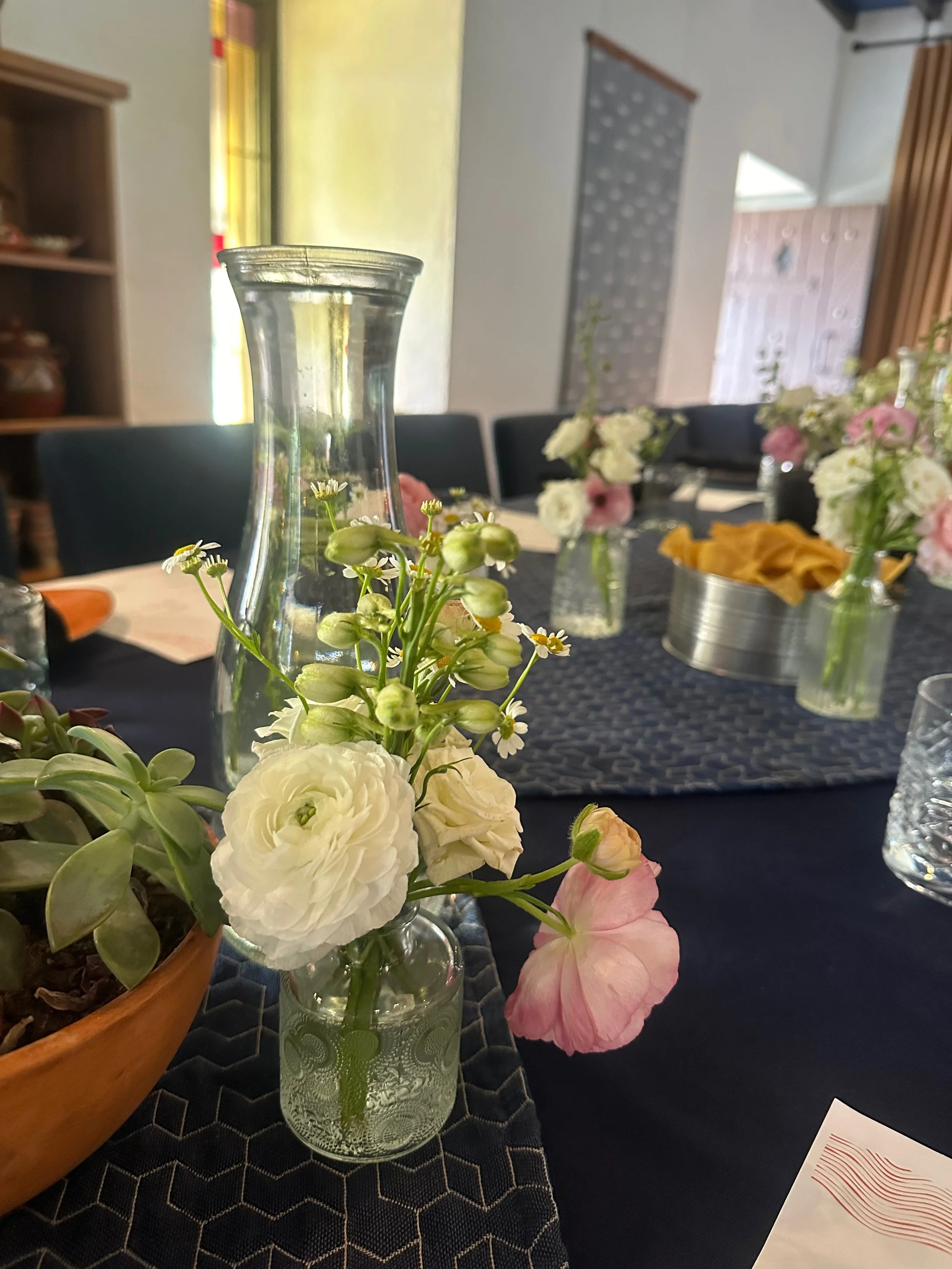 A dining table decorated with glass vases of white and pink flowers, including roses and ranunculus, with a dark table runner and chairs in the background.
