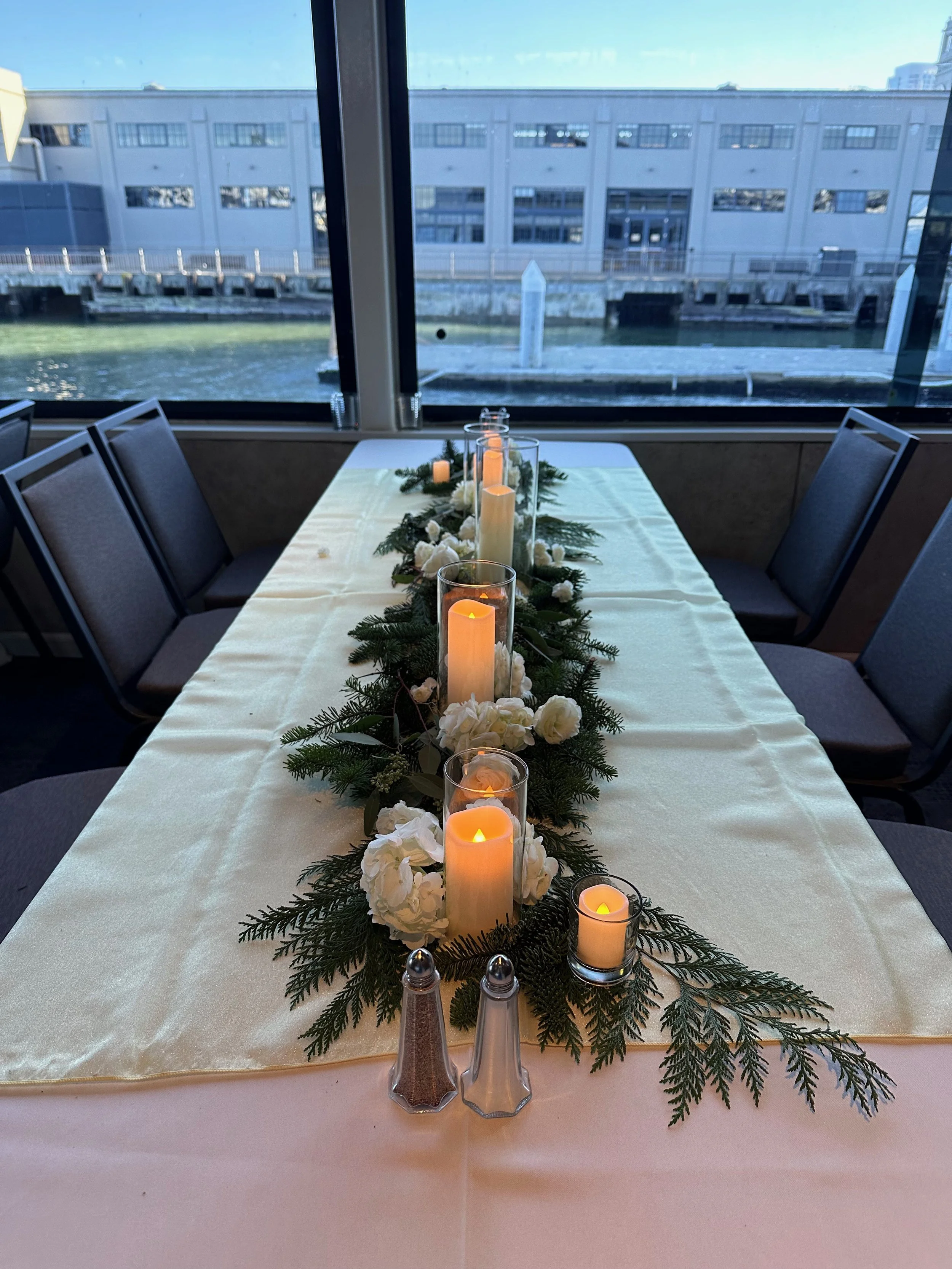 Table decorated for a festive occasion with a white tablecloth, candles in glass holders, green pine branches, and white flowers, positioned near large windows overlooking a waterfront building.