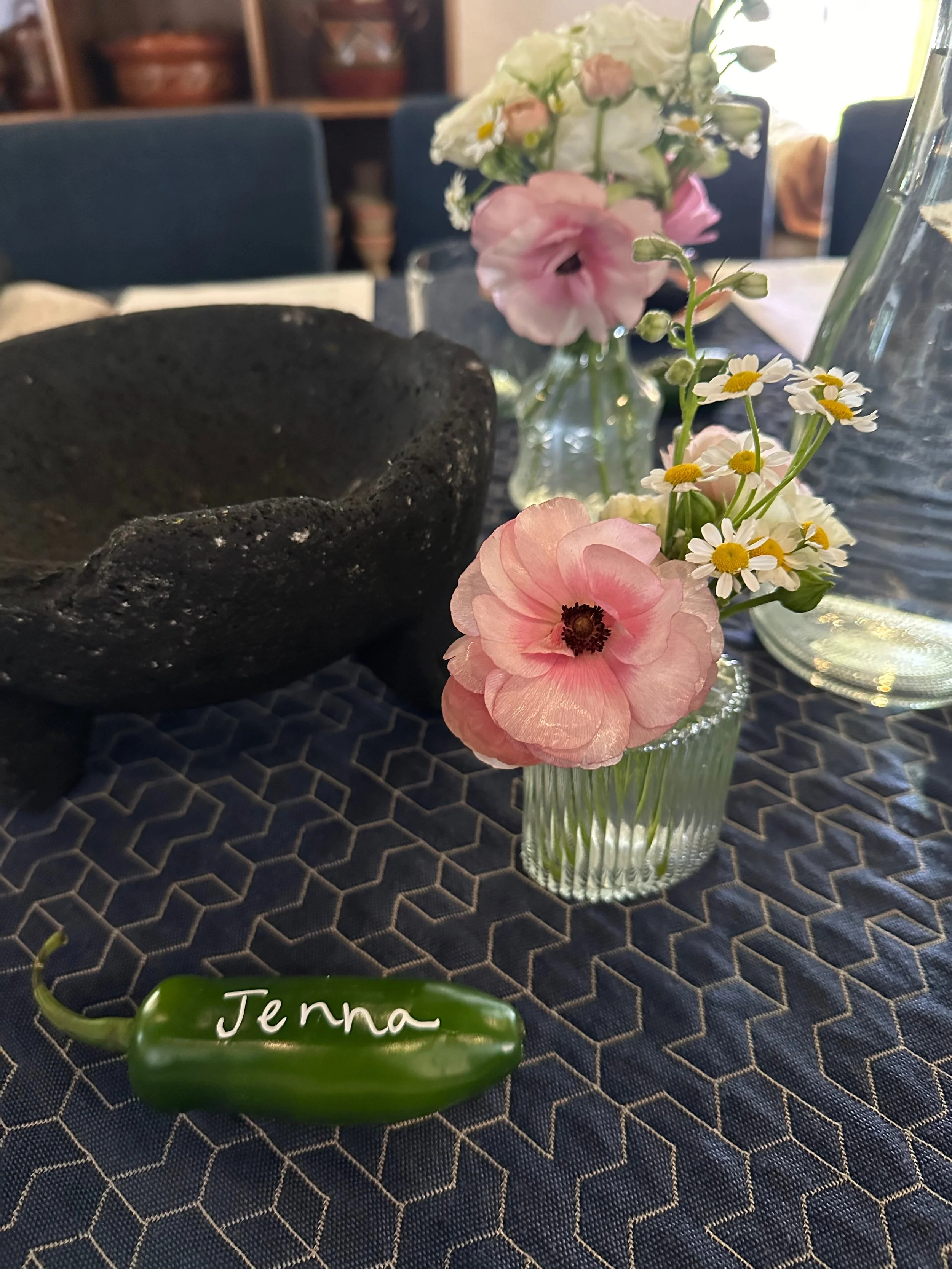 A small glass vase holding pink and white flowers on a patterned tablecloth. A green jalapeño pepper with the name 'Jenna' written on it rests nearby. In the background, a large black bowl and a larger glass vase are visible, with a dining room setti