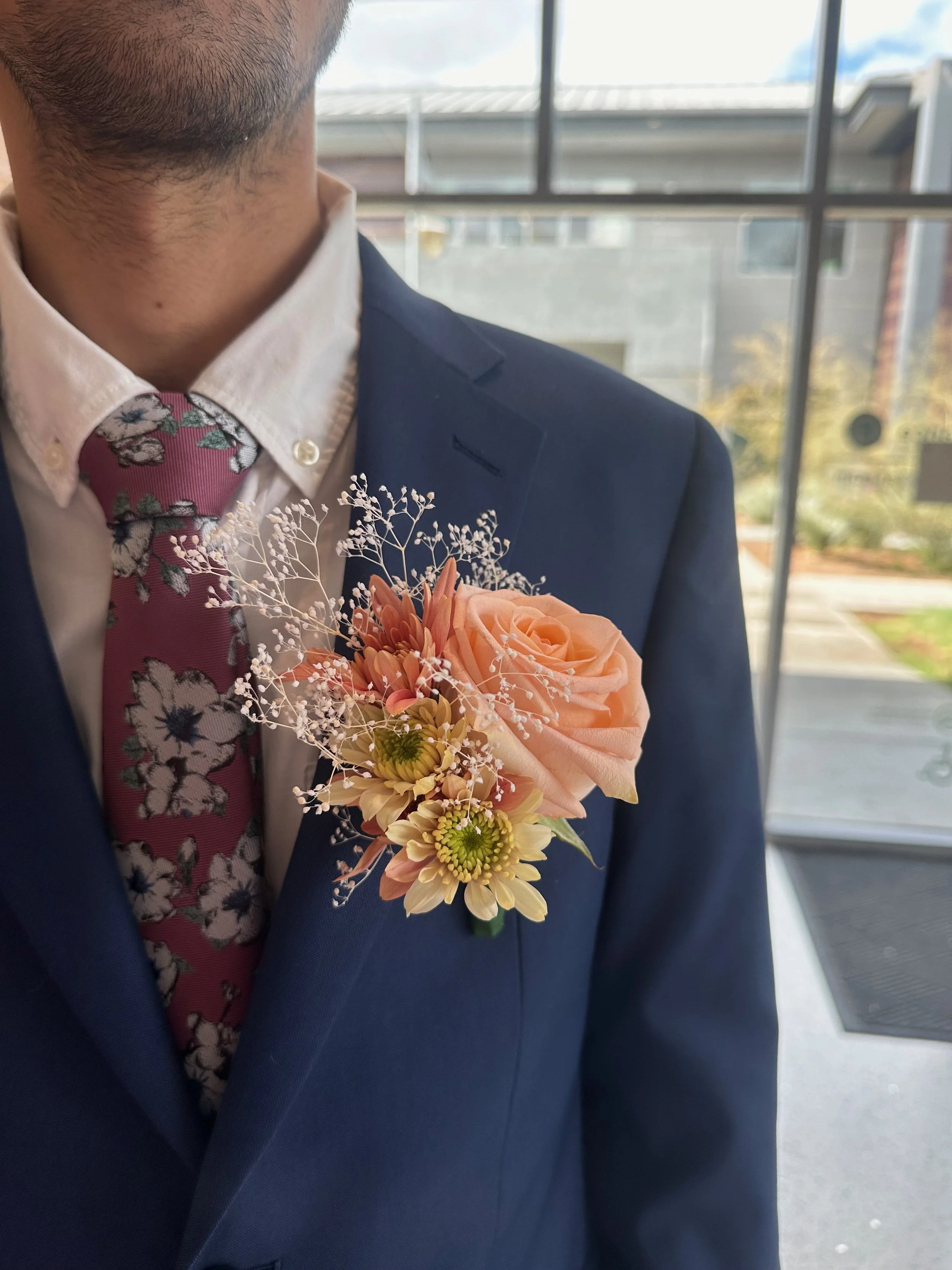 A man in a navy suit with a white shirt and floral patterned tie, wearing a peach-colored rose boutonniere on his chest, standing inside a building near large windows with an outdoor view.