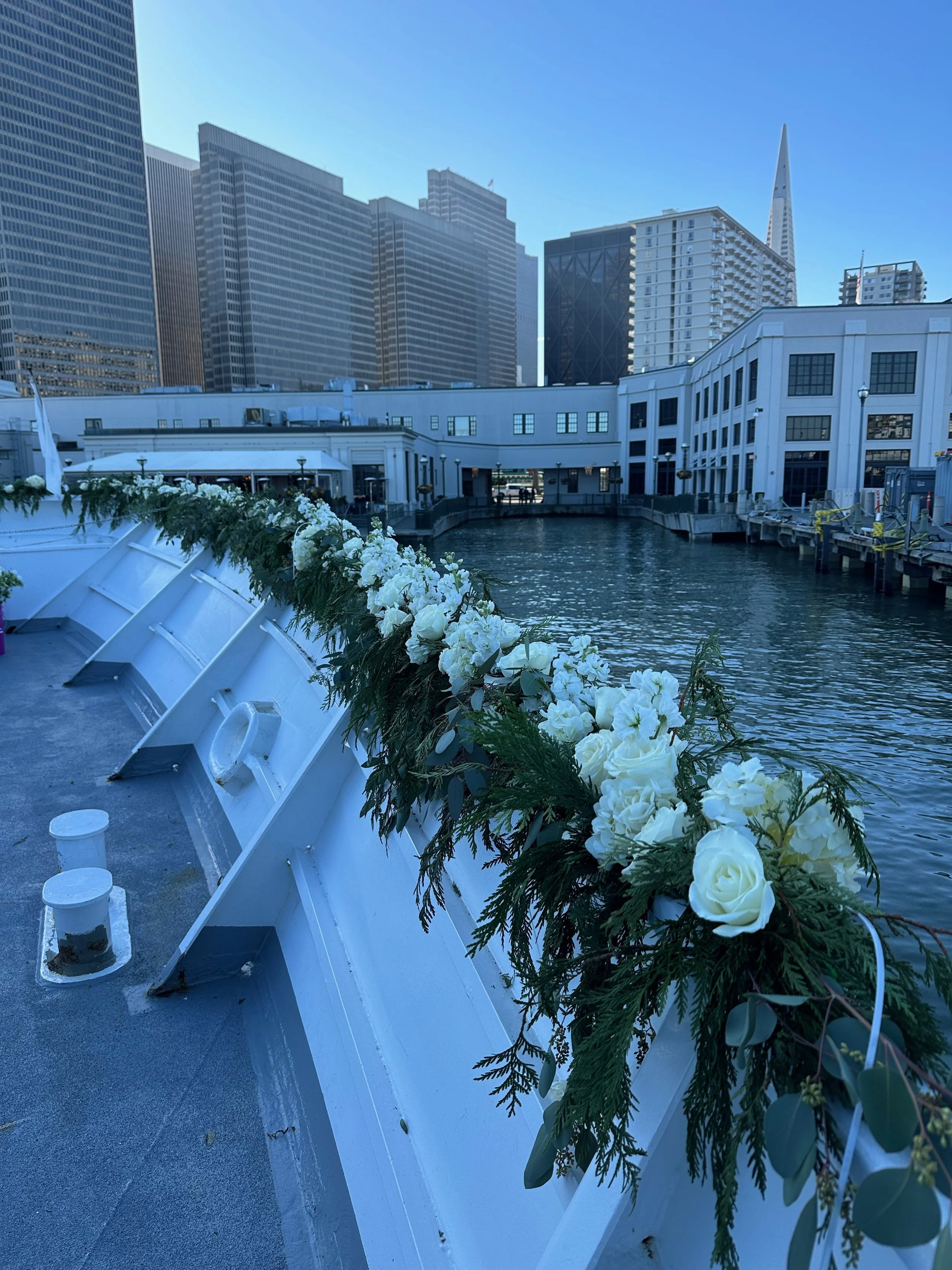 Decorative white floral arrangement along the railing of a docked boat on a waterway in an urban area with tall skyscrapers in the background.