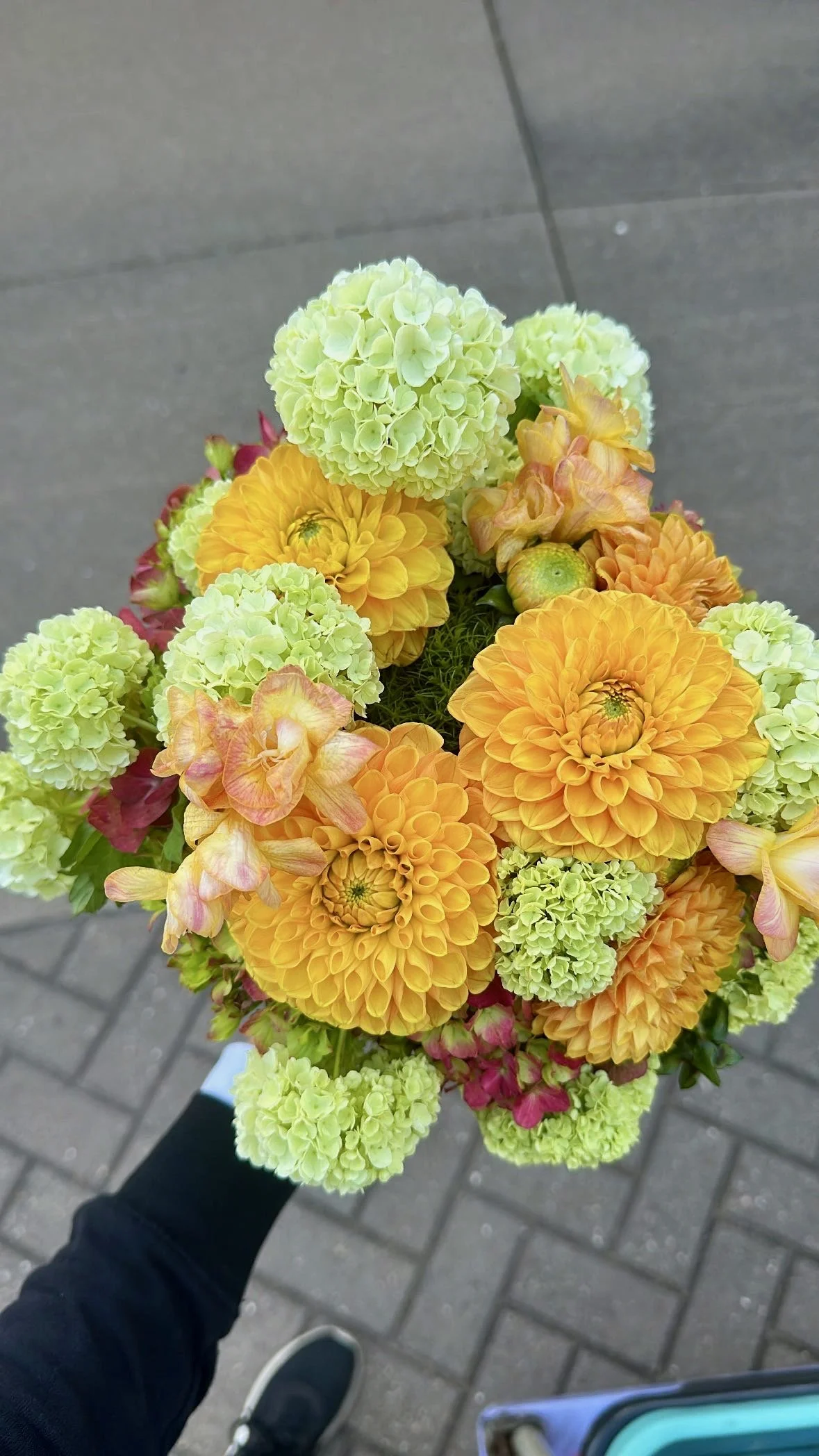 A person's hand holding a bouquet of yellow and green flowers, including dahlias and hydrangeas, against a pavement background.