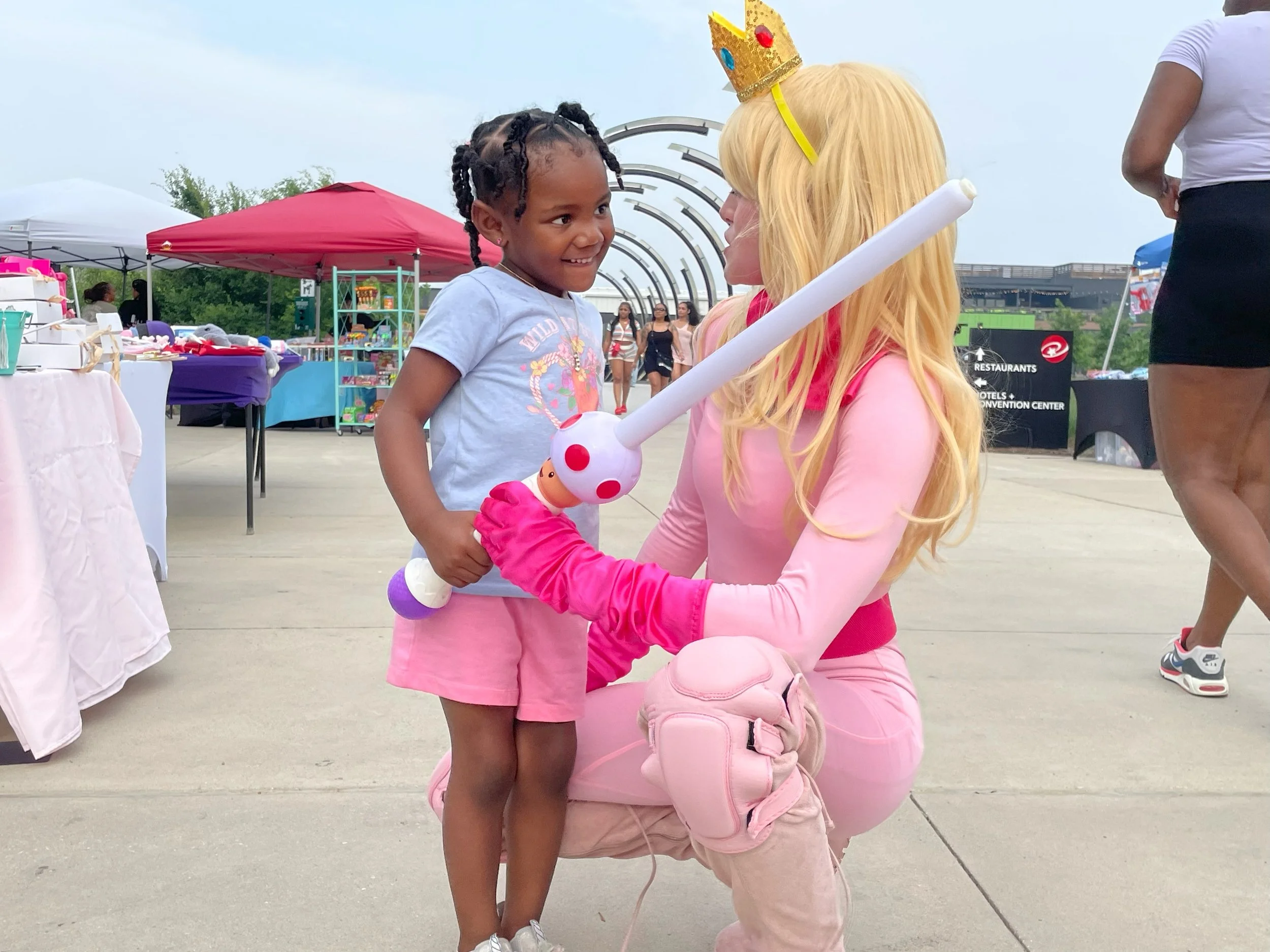A young girl smiling at a woman in a pink costume with blonde hair and a crown, at an outdoor event. The woman is holding a toy and squatting to interact with the child. Background includes event tents and people.