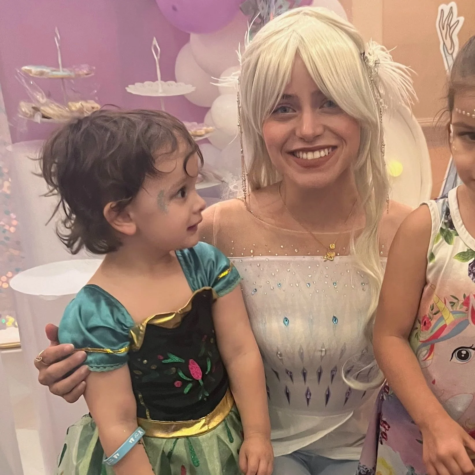 A woman dressed as a princess with two children at a party. The woman has long blonde hair and is wearing a white dress with blue rhinestones. One child is in a green costume, and the other child is in a colorful unicorn dress. The background includes white and purple balloons and dessert stands.
