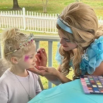 A woman dressed as a princess applies makeup to a young girl wearing a tiara.