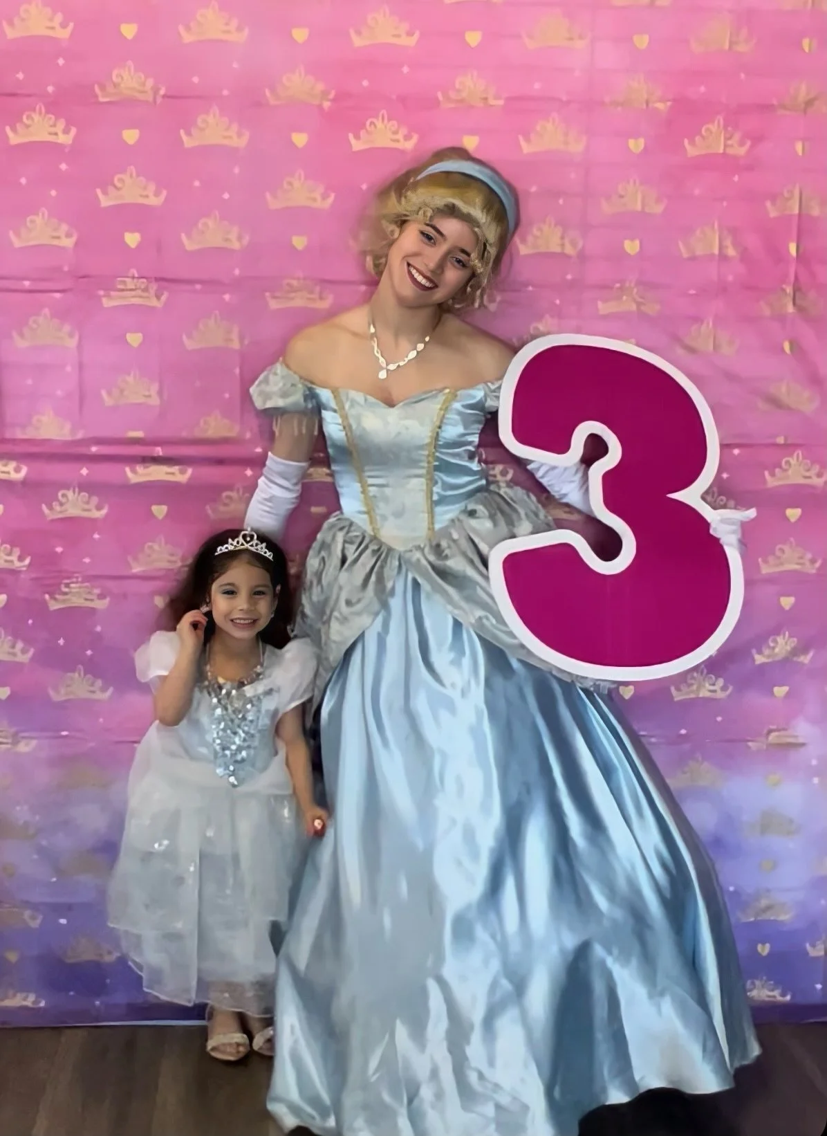 Woman dressed as a princess with a girl in a princess dress holding a number three sign, standing against a pink backdrop with crown patterns.