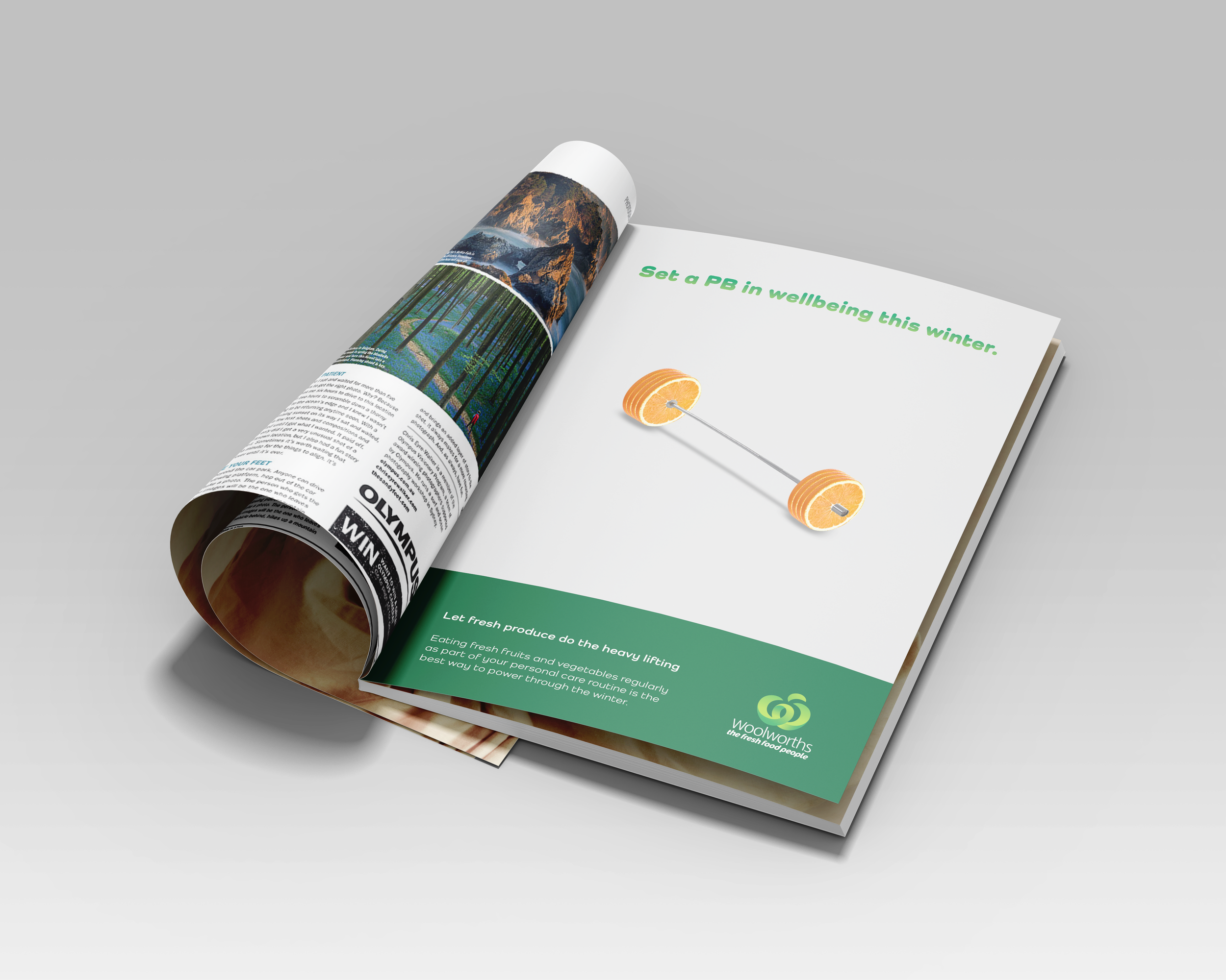 Open magazine with an article about wellbeing, showing a colorful landscape photo of a lake and trees and an advertisement for fruit-based exercise equipment that resembles weights with oranges, on a grey surface.