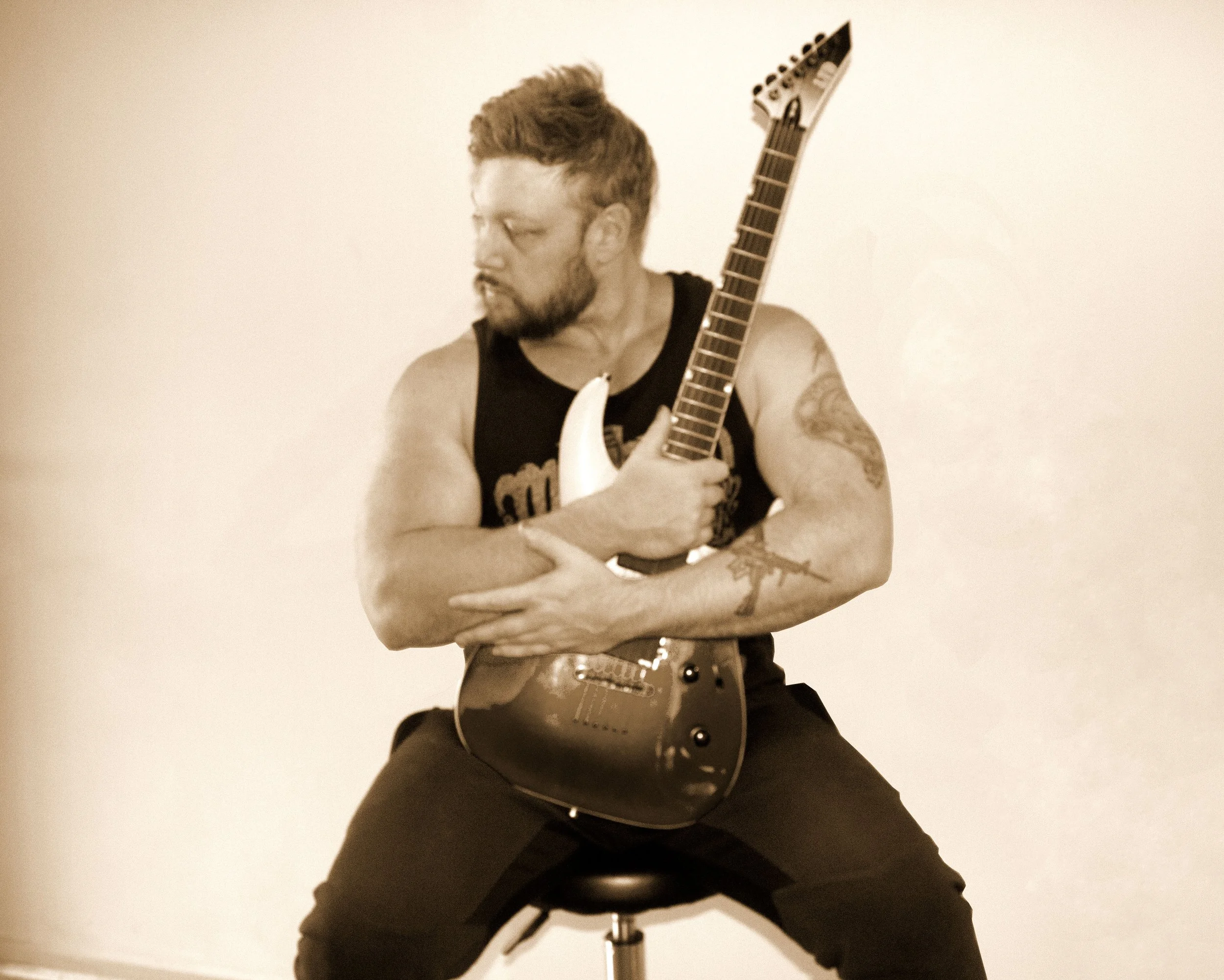 A man with tattoos on his arms, wearing a black sleeveless shirt, sitting on a stool while holding an electric guitar against a plain wall.