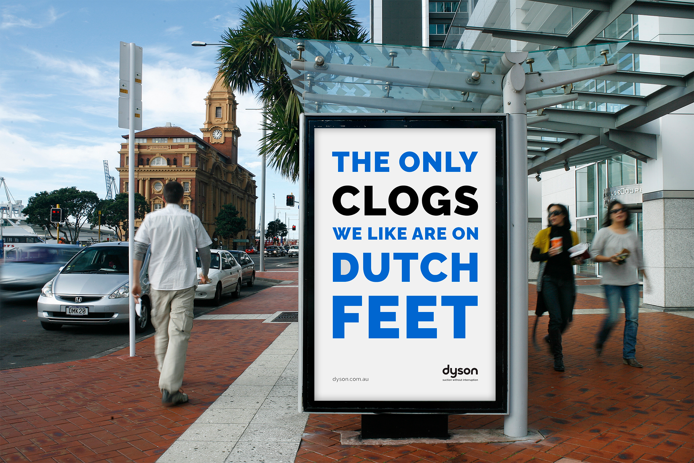 Urban street scene with pedestrians, cars, and a large bus stop advertisement that reads, 'The only clogs we like are on Dutch feet,' with a logo for Dyson at the bottom.