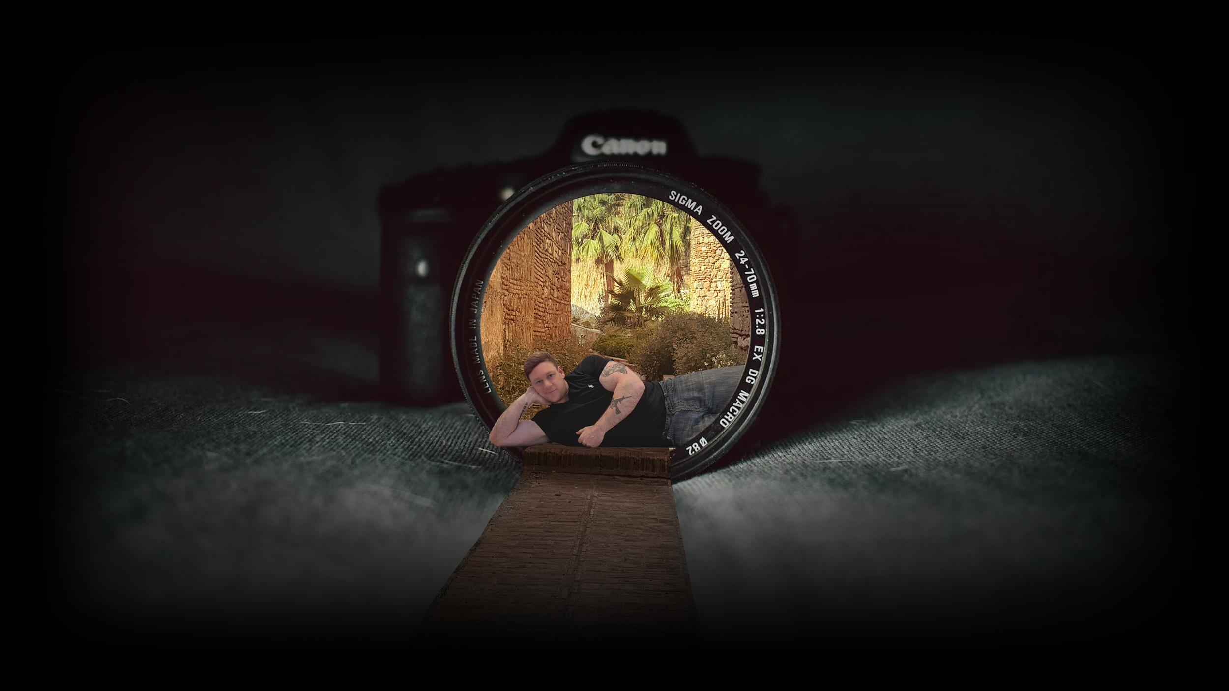 A digital photo of a person lying on a wooden walkway, with their head propped up on one arm, inside a camera lens, with a background of outdoor plants and trees.