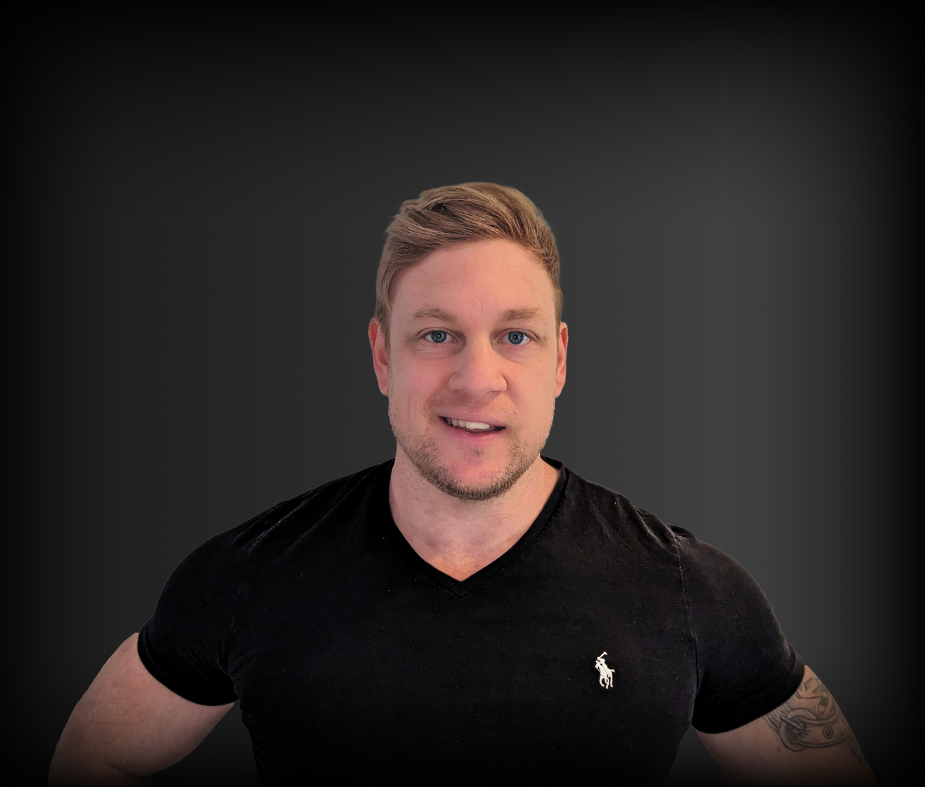 A young man with short, blonde hair, blue eyes, and light facial hair wearing a black v-neck t-shirt with a small white logo on the chest, standing against a dark gray background.