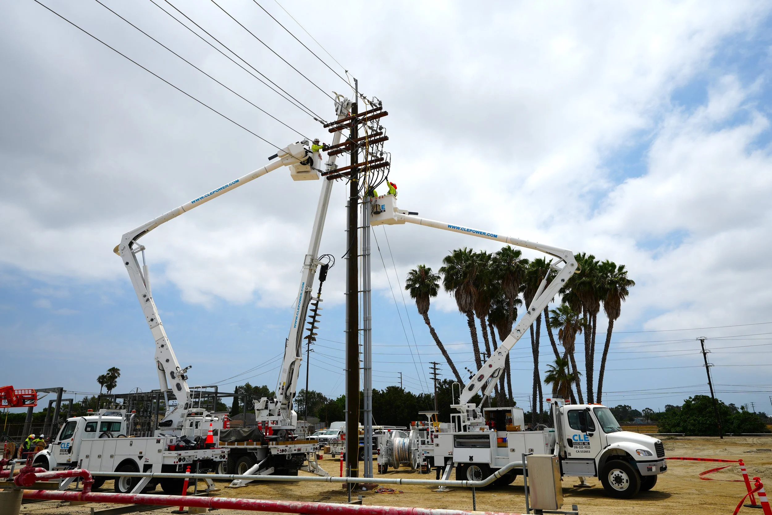Workers repairing or maintaining electrical power lines using bucket trucks at a utility pole in an outdoor area with palm trees.