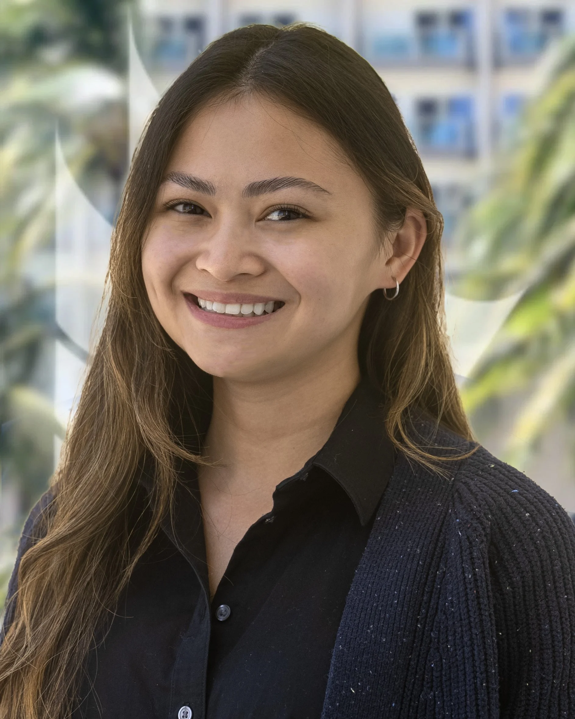A smiling woman with long brown hair, wearing a black shirt and a dark sweater, in front of a blurred background with greenery and buildings.