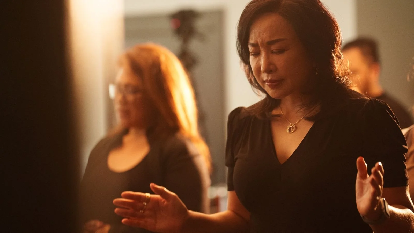 A woman with dark hair and eyes closed, wearing a black dress and a gold necklace, appears to be praying or deep in thought, with her hands raised and eyes closed, in a dimly lit room.