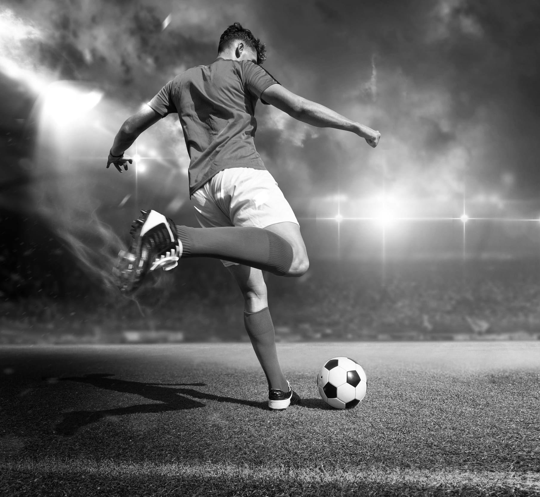 A soccer player kicking a ball on a field with dramatic sky and stadium lights in the background, black and white image.