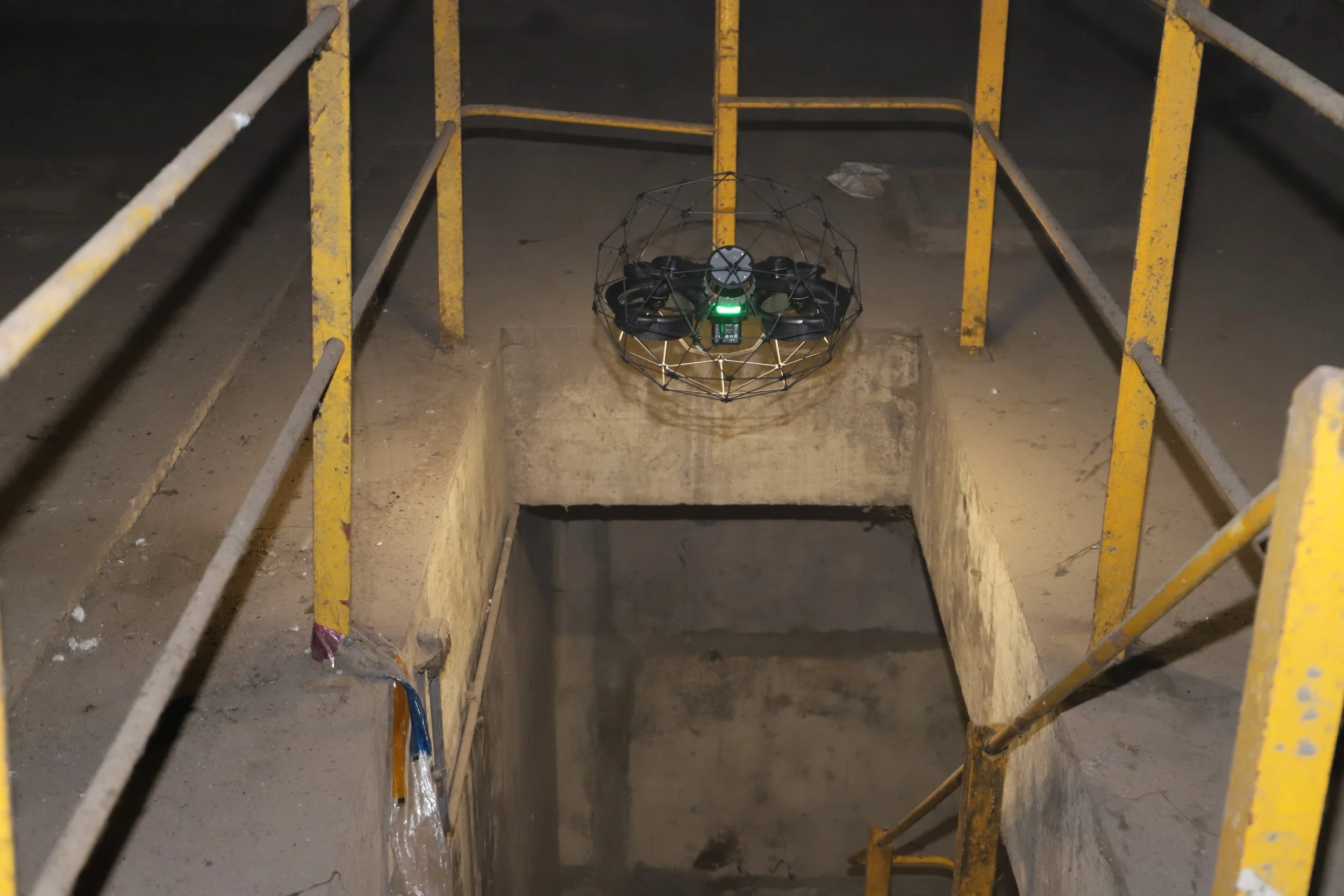 A confined space drone with a protective cage hovers near a construction or maintenance opening in the ground, surrounded by yellow railings in Hong Kong.