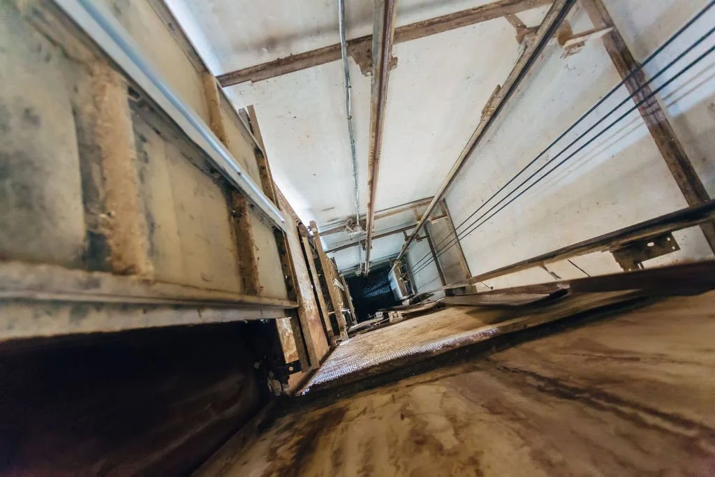 Looking down an elevator shaft showing metal support beams, cables, and the elevator car at the bottom, with the perspective from near the top using a confined space inspection drone.