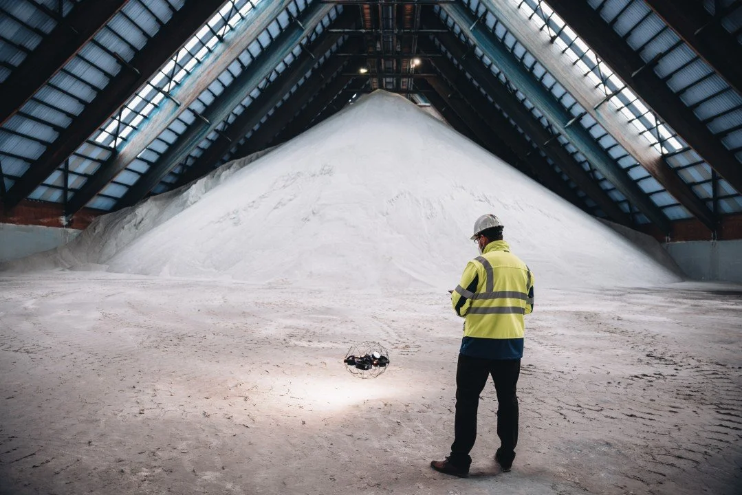 A worker in a yellow safety jacket and helmet operating a drone in a large warehouse filled with a mountains of salt or similar white mineral in confined space using a drone for inspection.