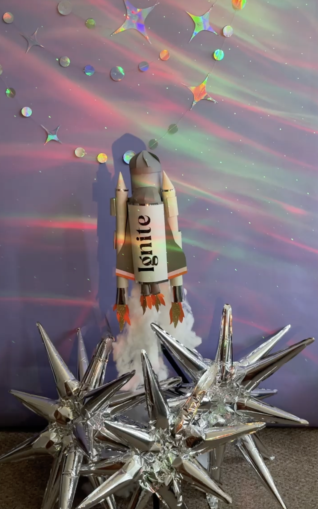 A toy rocket labeled 'light' is launching into a starry, colorful background with holographic star-shaped decorations and shiny silver star-shaped objects on the ground.