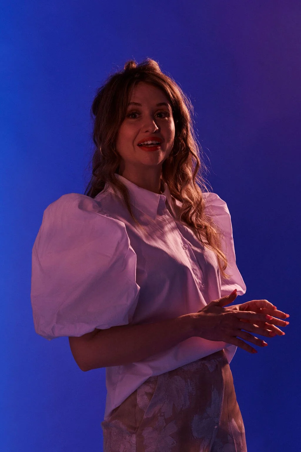 A woman with long wavy hair in a pink blouse with puffed sleeves stands against a blue-purple background.