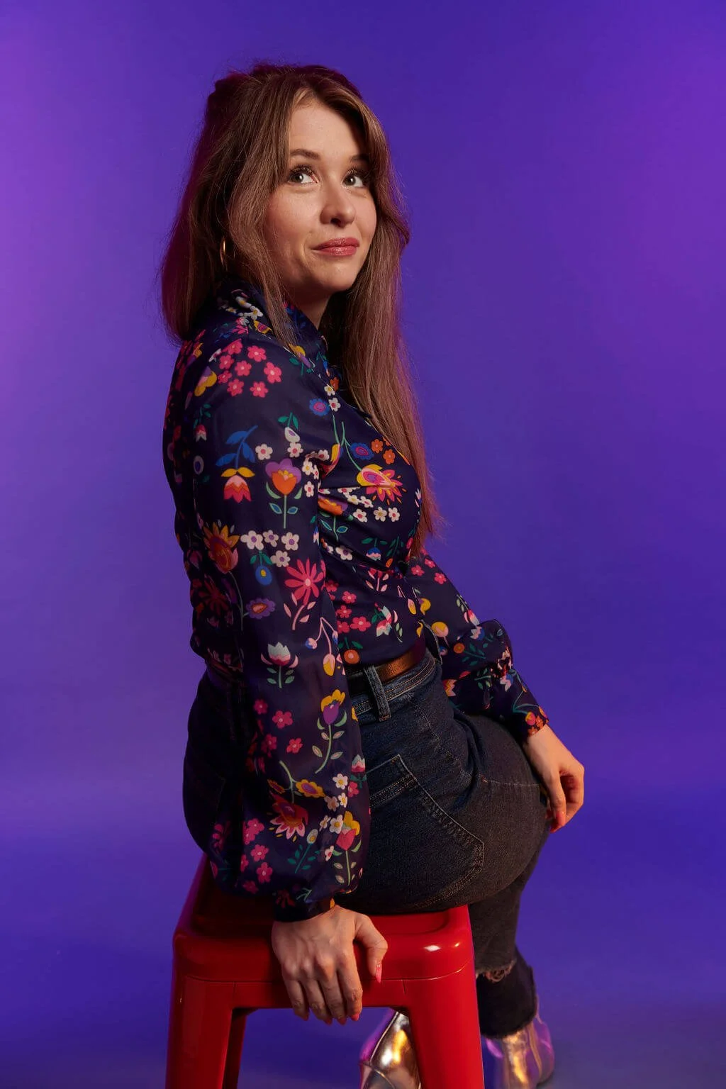 Woman with long brown hair sitting on a red stool against a purple background, wearing a colorful floral blouse and dark jeans.