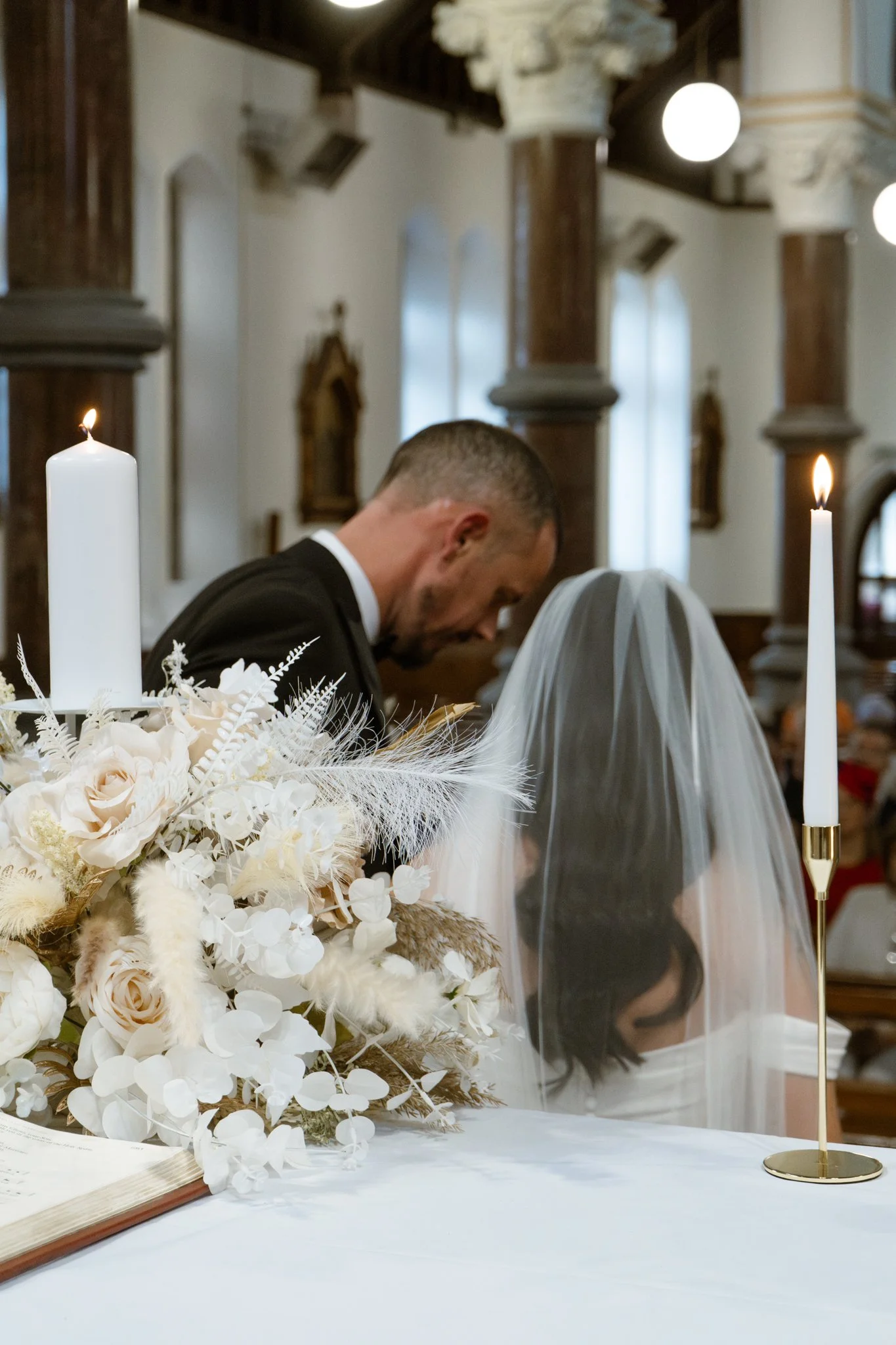 A bride and groom kneeling and praying during a wedding ceremony inside a church, with candles and a bouquet of white flowers on the table in the foreground.