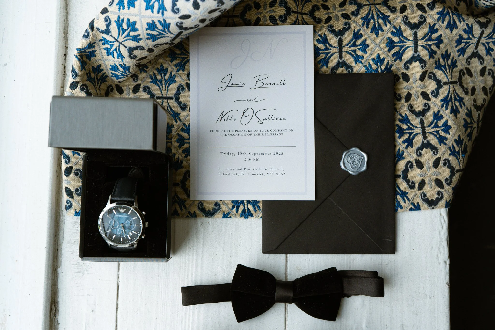 Wedding invitation, wristwatch in a box, black envelope with wax seal, black bow tie, floral patterned fabric in background.
