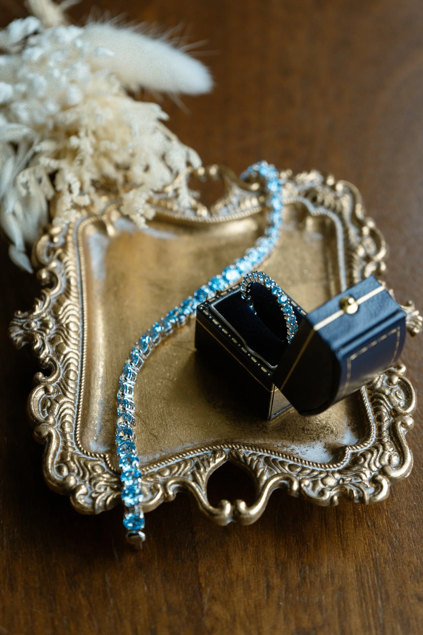Jewelry and accessories including a diamond necklace, bracelet, and ring displayed on an ornate silver tray, with a black jewelry box and white floral decoration in the background.