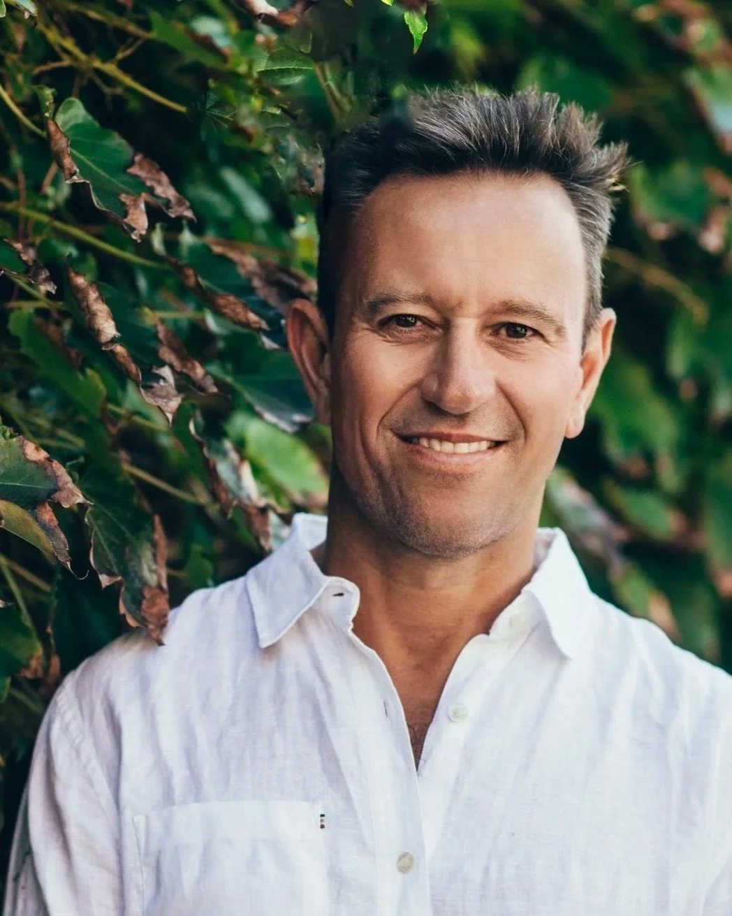 Smiling man in a white shirt stands in front of leafy green plants, with sunlight highlighting his face.