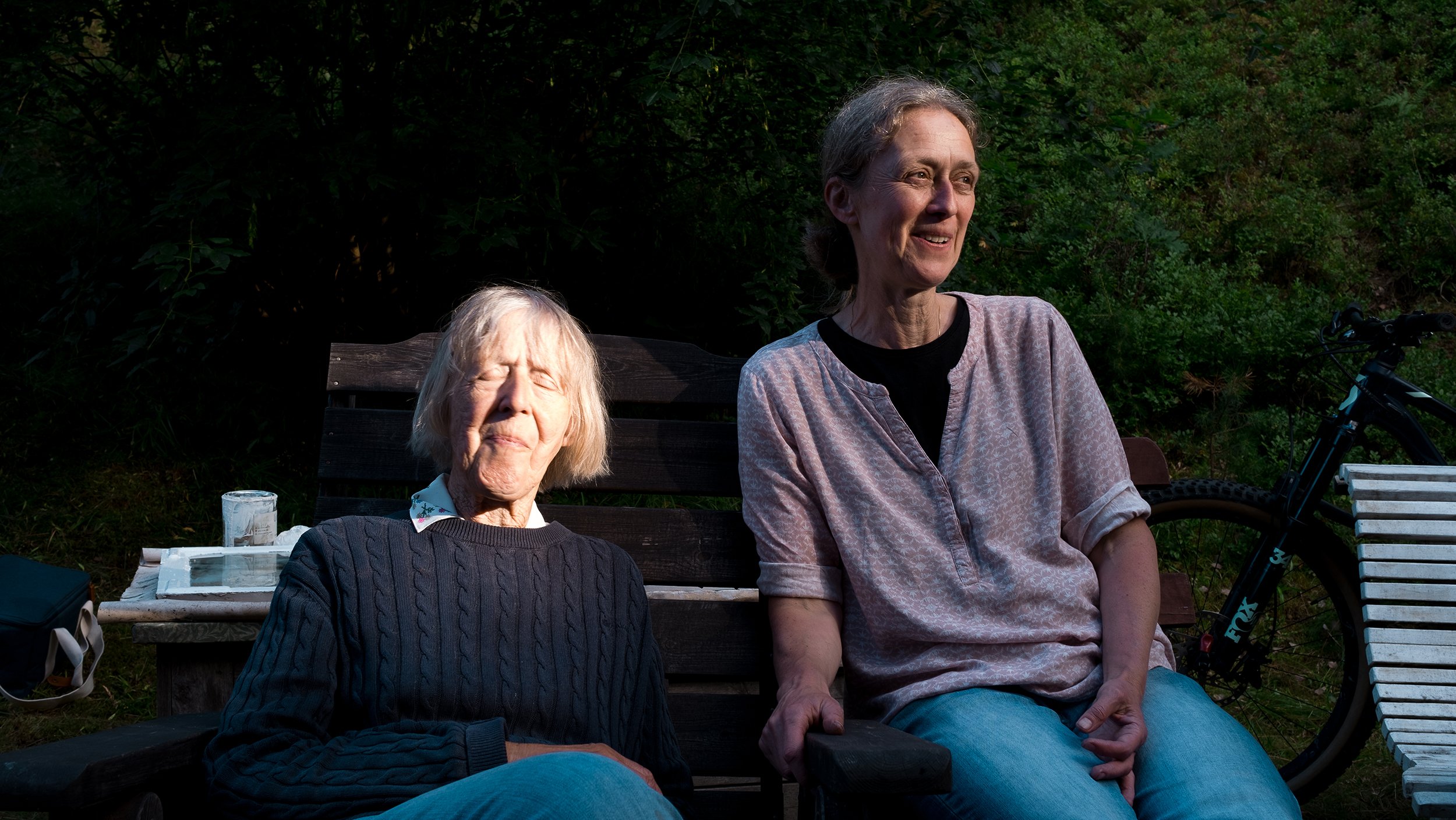 Two women sitting on a wooden bench outdoors, with a bicycle partially visible on the right. One woman is wearing a dark sweater and has her eyes closed, while the other is wearing a patterned shirt and smiling. There is green foliage in the backgrou