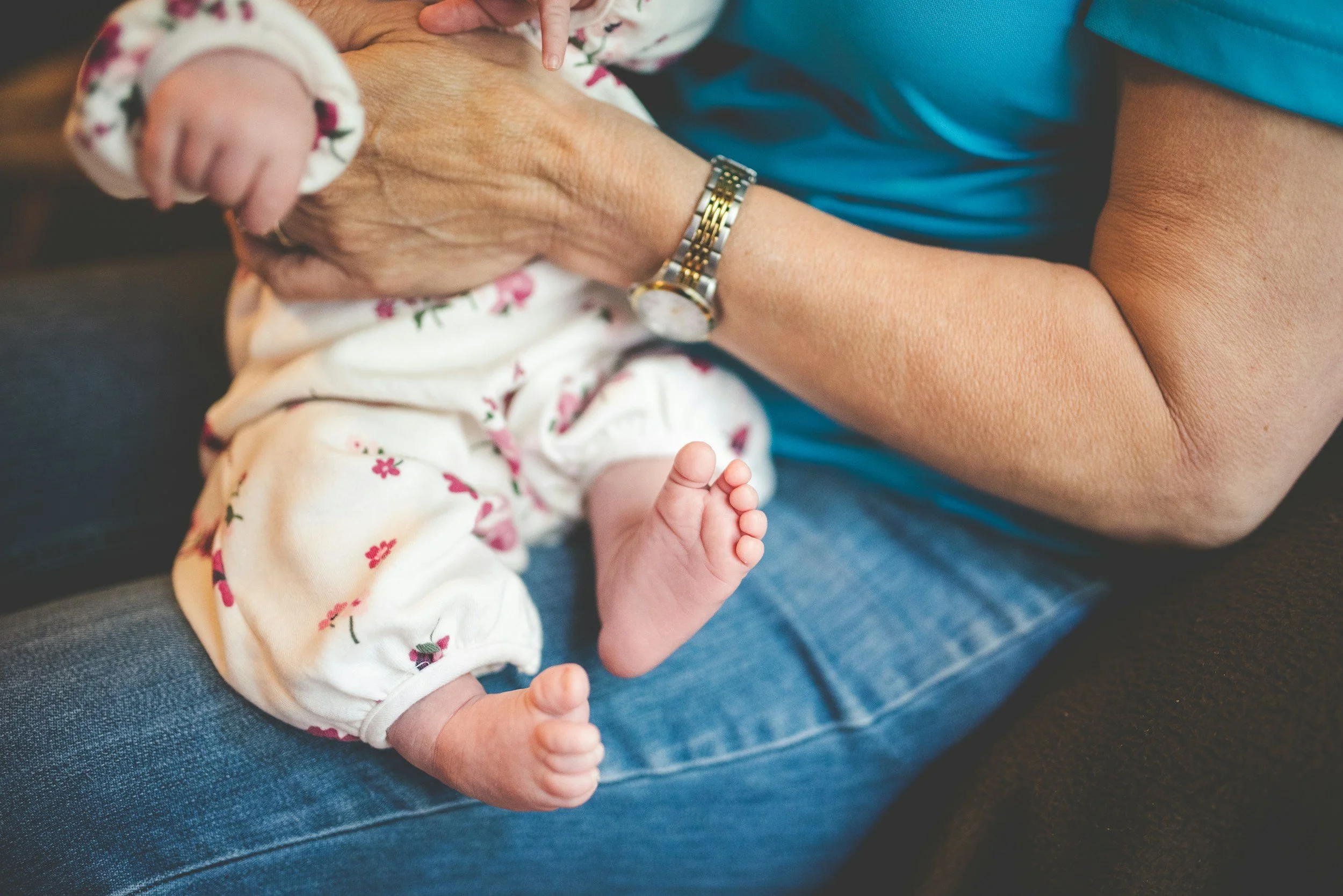 A baby sitting on an adult's lap, with the adult wearing a watch and blue jeans. The baby is wearing a floral long sleeve top and has tiny feet, with one foot slightly raised.