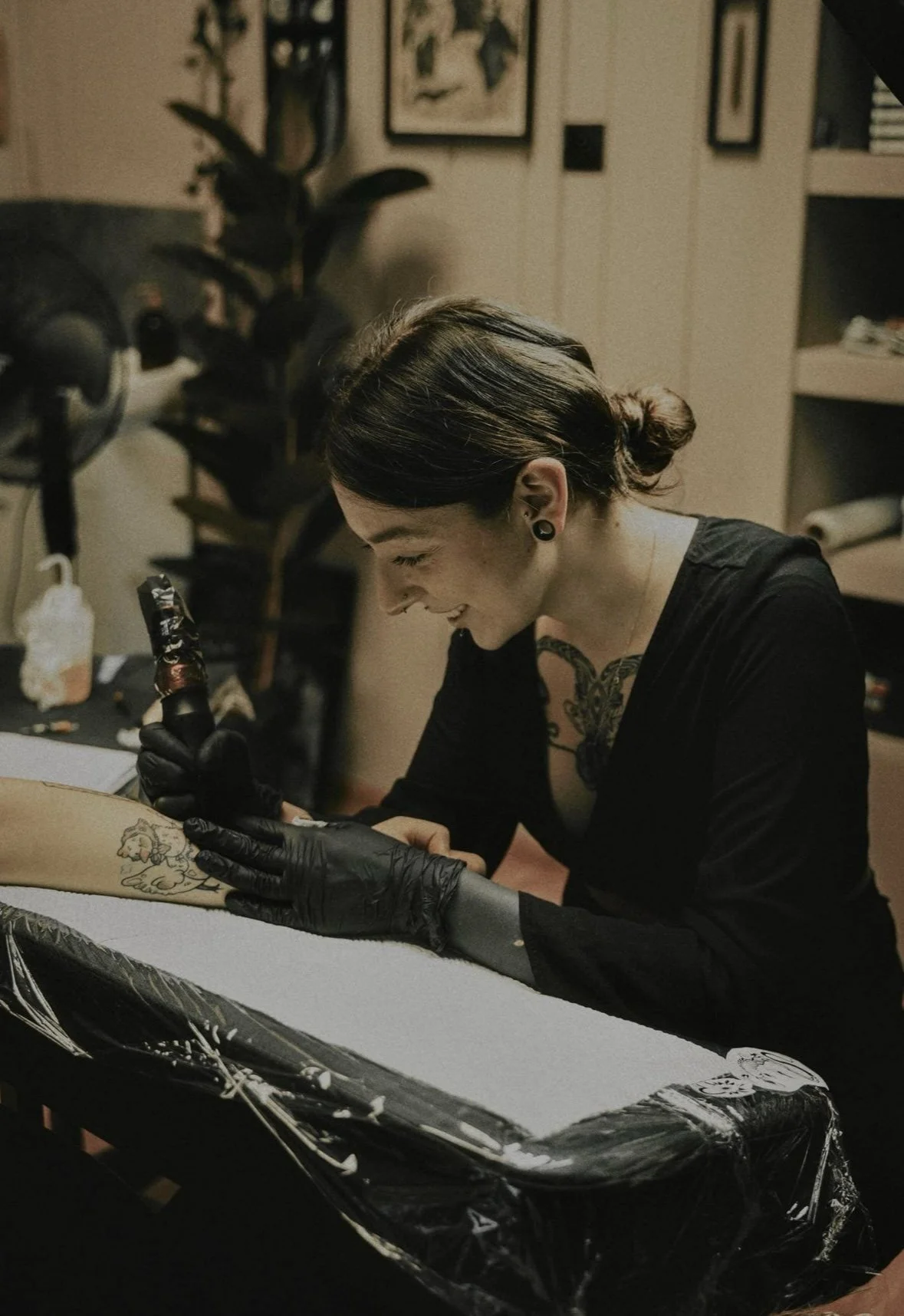 A woman with dark hair in a bun, wearing black gloves and earrings, is tattooing a lion on another person's arm in a workshop room. She is smiling and focused on her work.