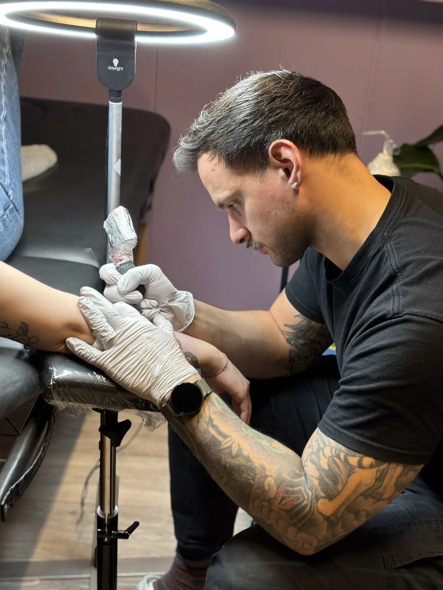 Tattooed man with earrings getting a tattoo on a person's ankle, with a tattoo machine in hand, under a bright ring light in a tattoo studio.