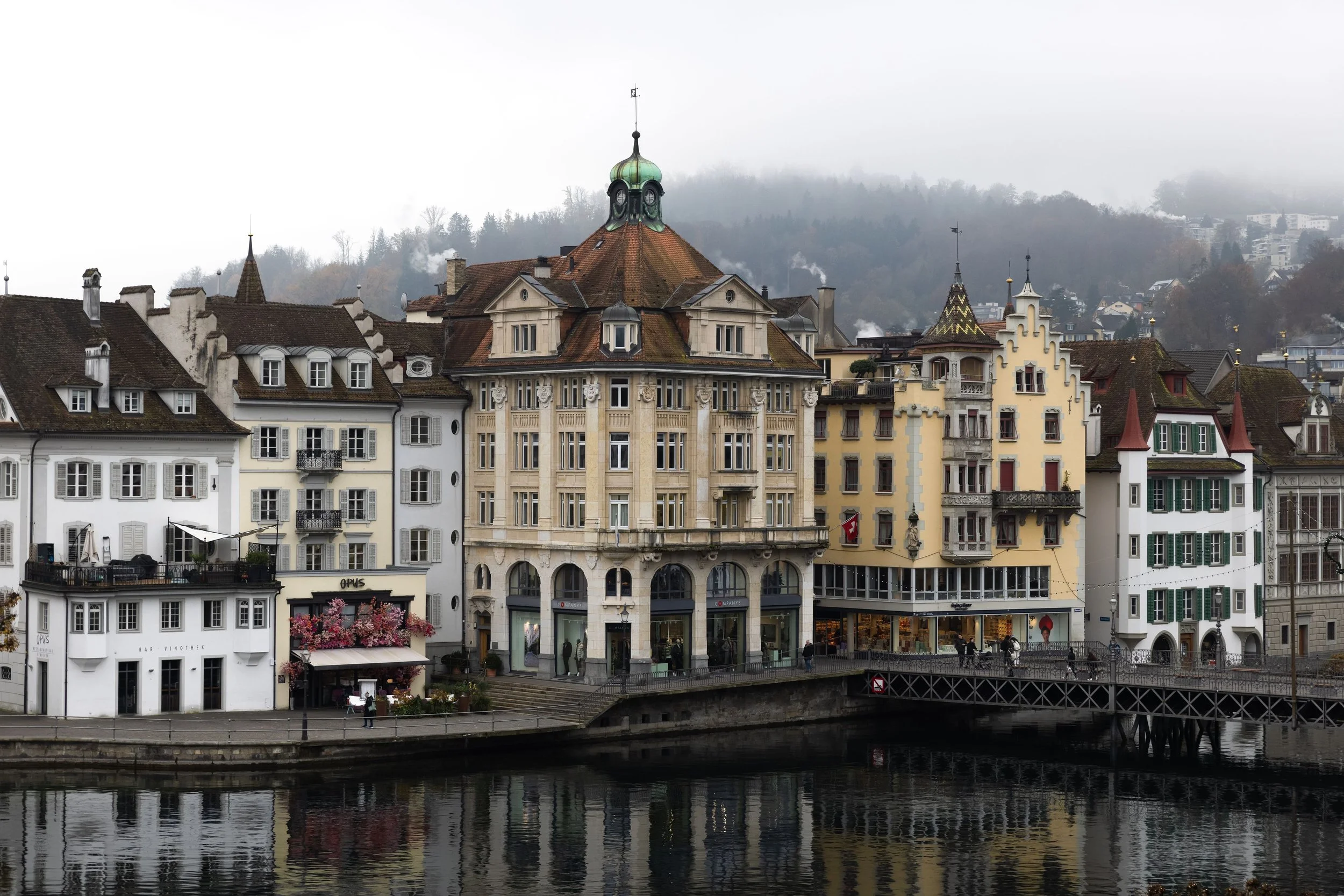 Cityscape of historic European buildings along a river, with overcast sky and misty hills in the background.