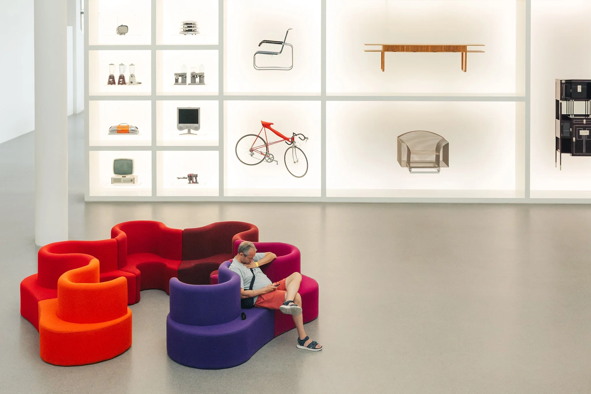 An indoor art gallery with a white grid wall displaying vintage electronic and technological devices, including an old bicycle, a small television, a stereo cabinet, and various other appliances. In the foreground, a man and a child are sitting on colorful, modern, rounded chairs, with the man using a smartphone.