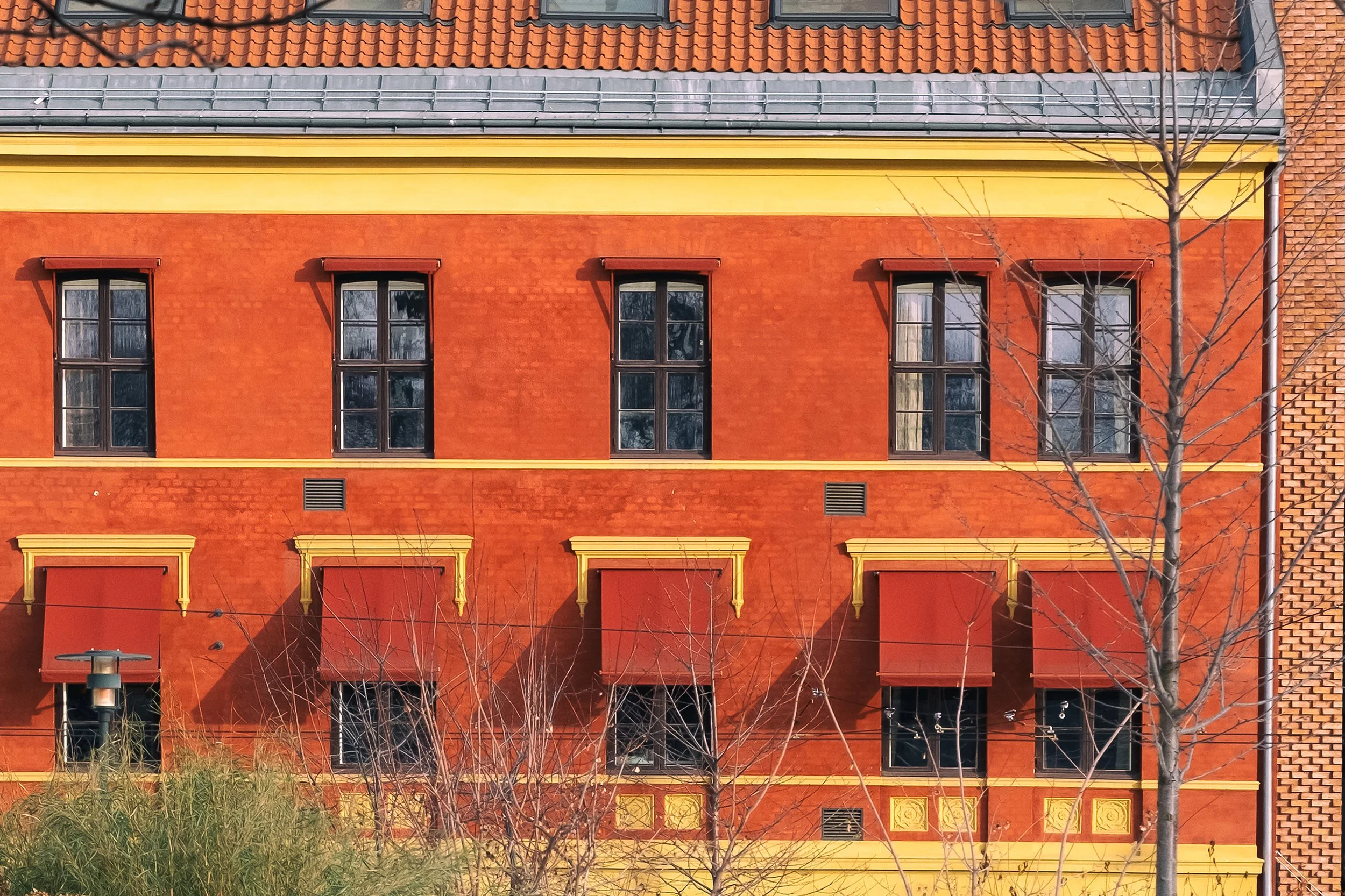 A colorful building with a red brick facade, yellow trim, and a tiled roof. There are three tall windows on the upper floor and a tree with bare branches in front.