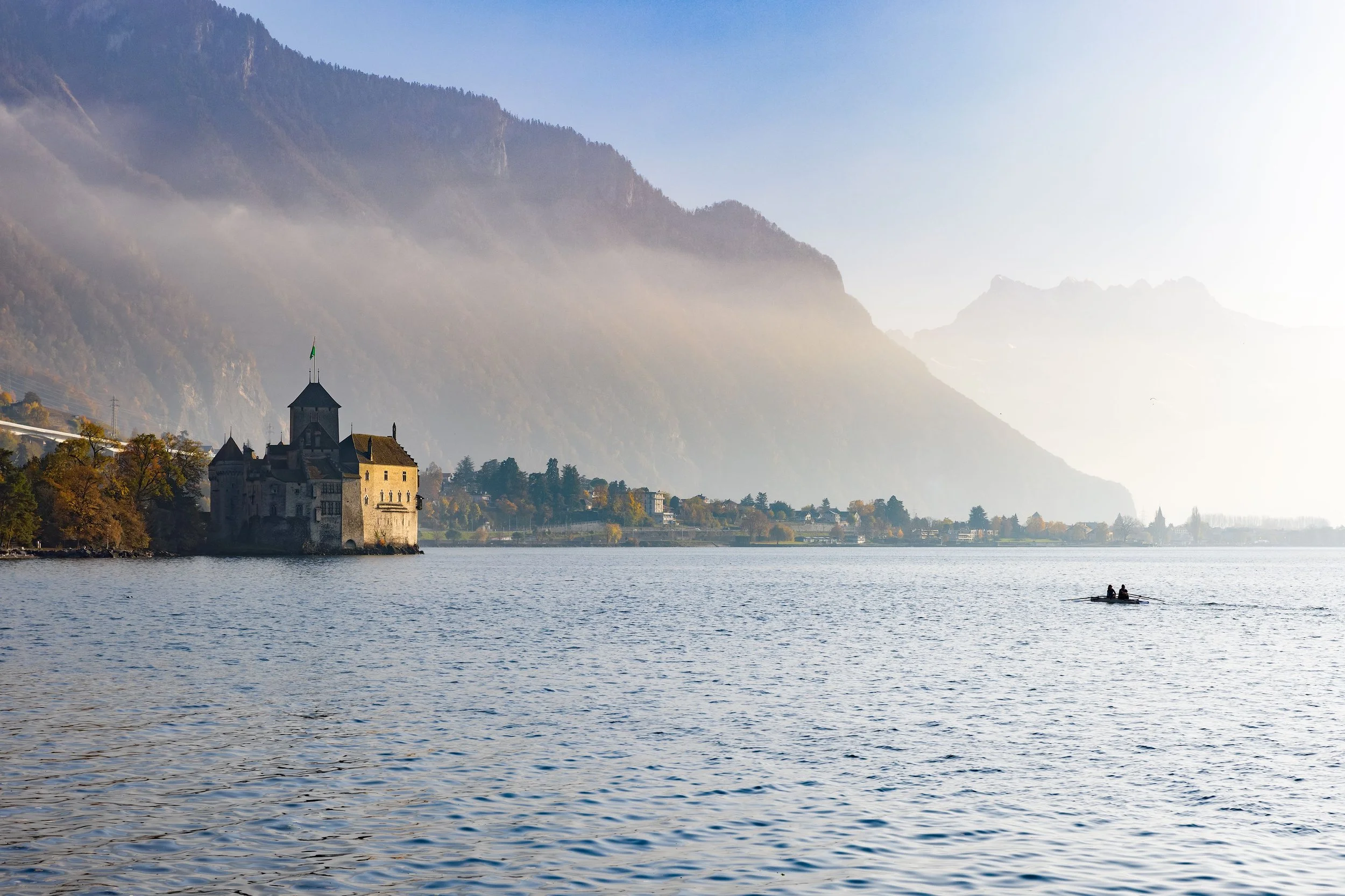 A calm lake with two people rowing a boat, surrounded by trees and mountains in the background, with mist or fog near the mountain base.