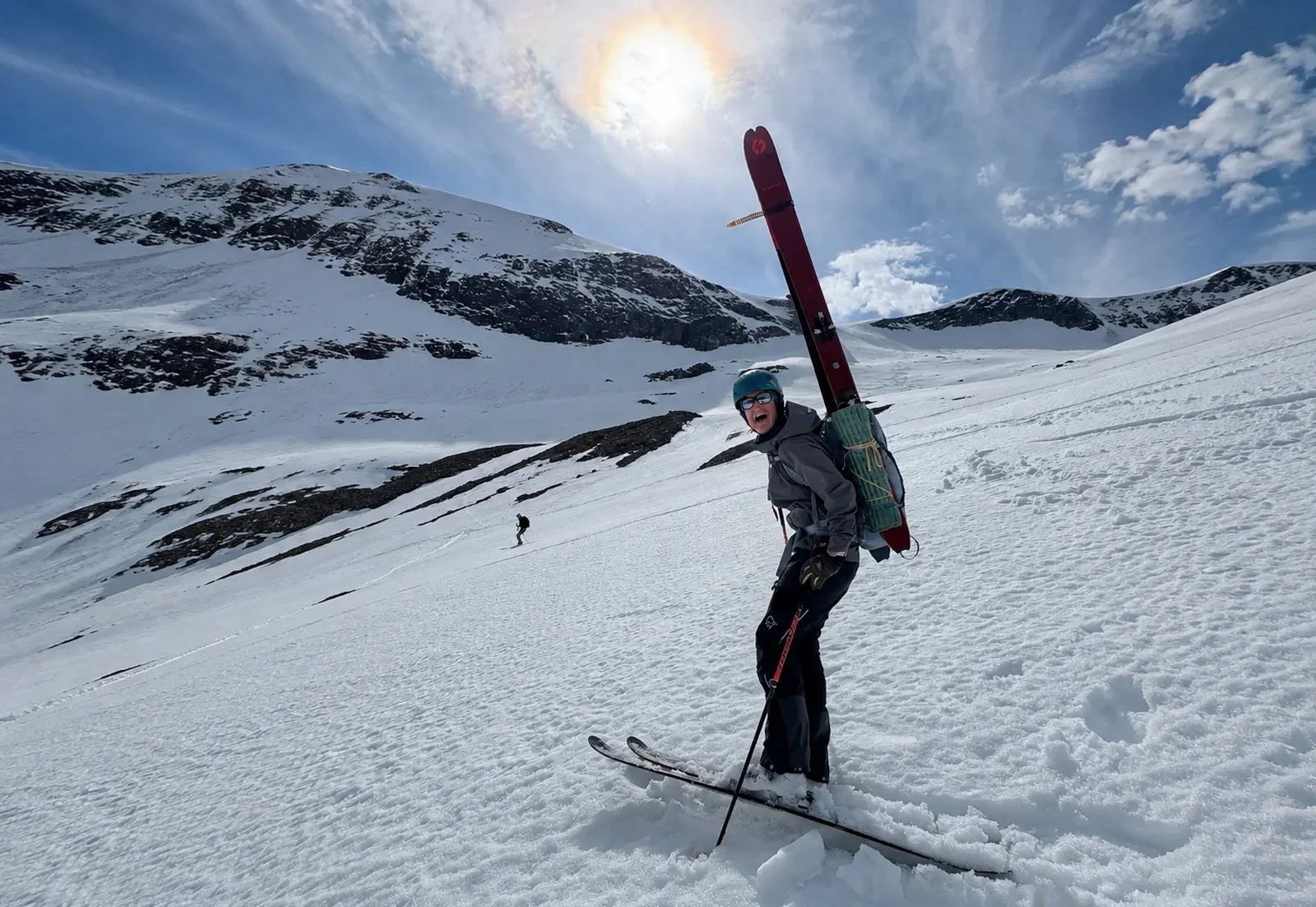A person wearing a helmet and sunglasses skiing on a snowy mountain slope, carrying skis on their back, with a bright sky and mountain landscape in the background.