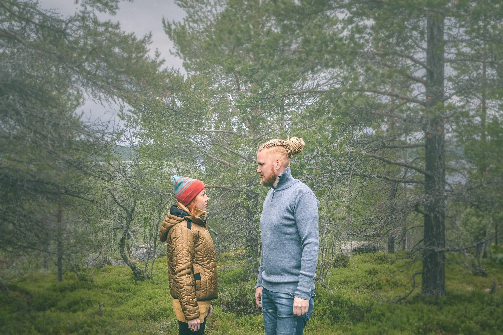 A man and woman standing face-to-face in a forested area with green trees and bushes around them, gazing into each other's eyes.