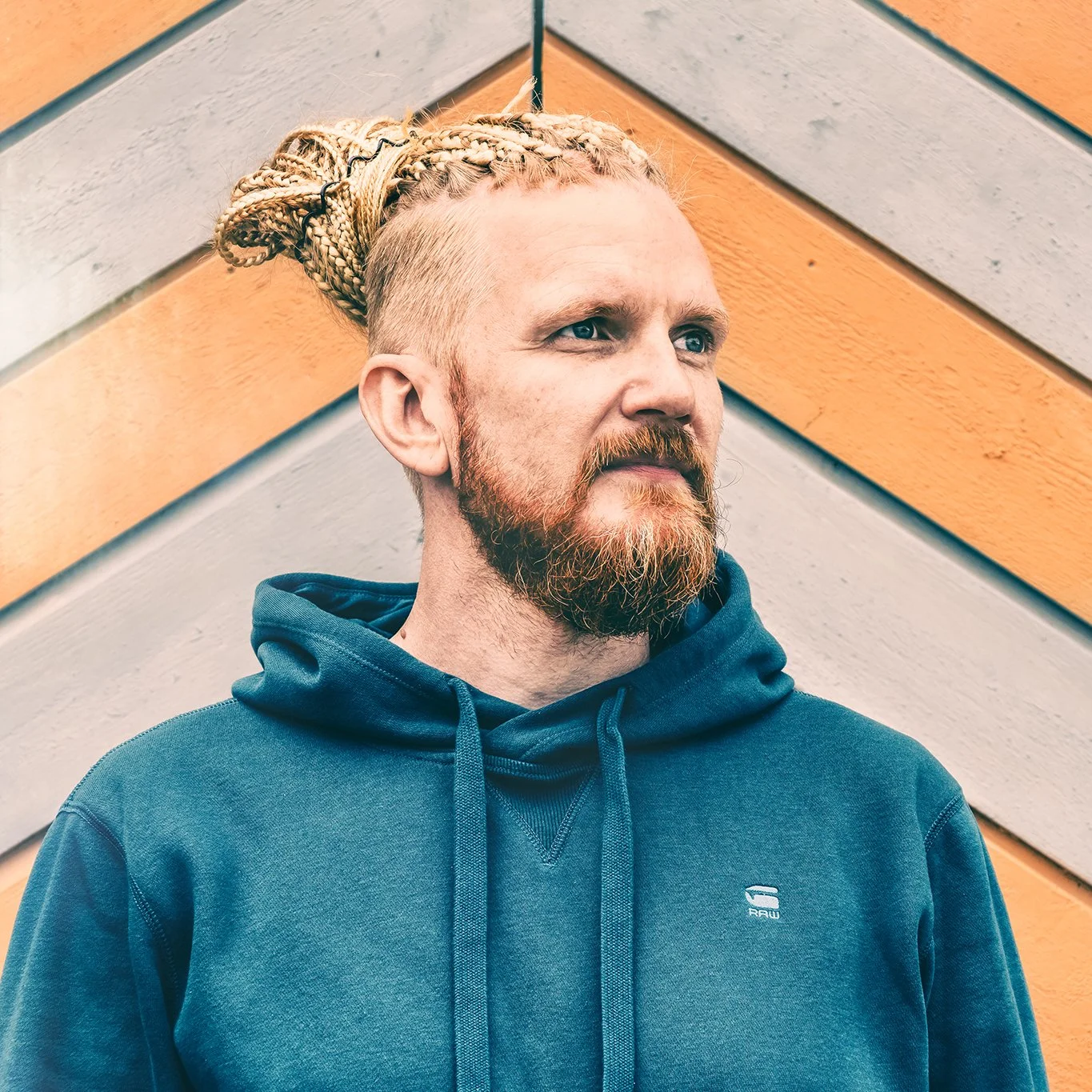A man with blond braided hair and a reddish beard stands in front of a diagonally striped orange and gray wall, wearing a blue hoodie.