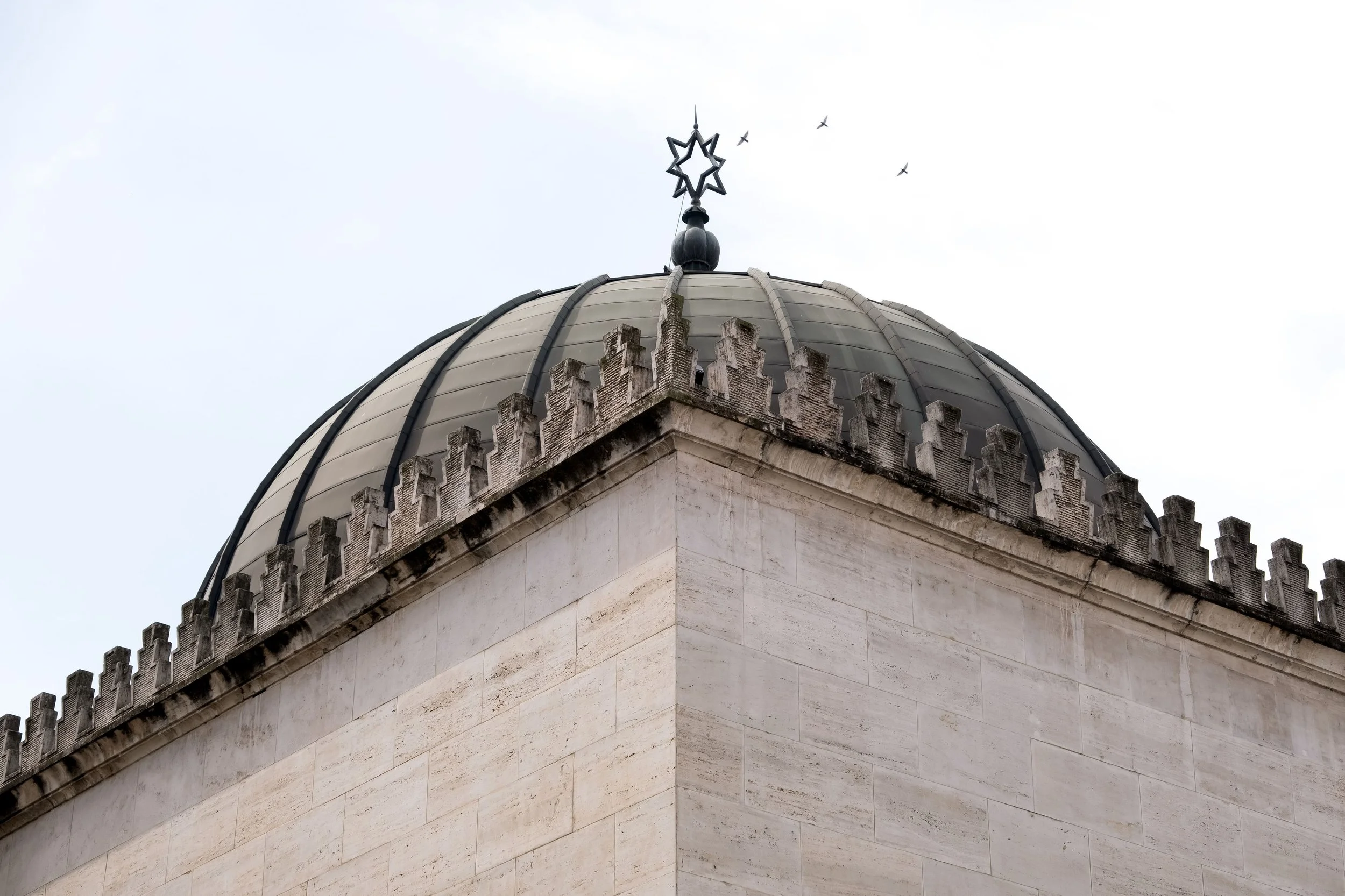 Close-up of the top part of a historic building with a dome and decorative medieval-style battlements, with birds flying in the cloudy sky.