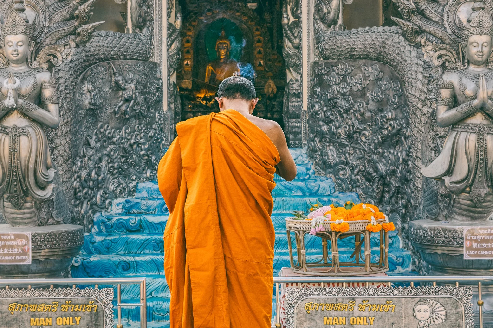 A Buddhist monk dressed in orange robes standing with back to camera inside a temple, facing a large ornate religious statue, with signs reading "MAN ONLY" and 'LADY CANNOT ENTER' at the front.