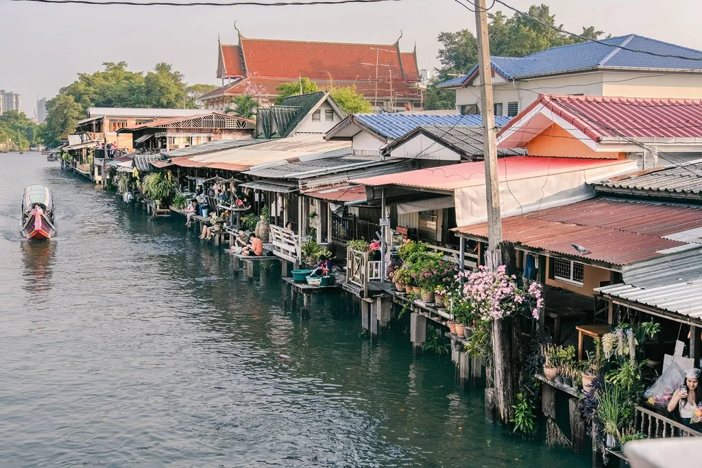 Waterside homes with colorful rooftops, boats on water, and people sitting outside on stilts.