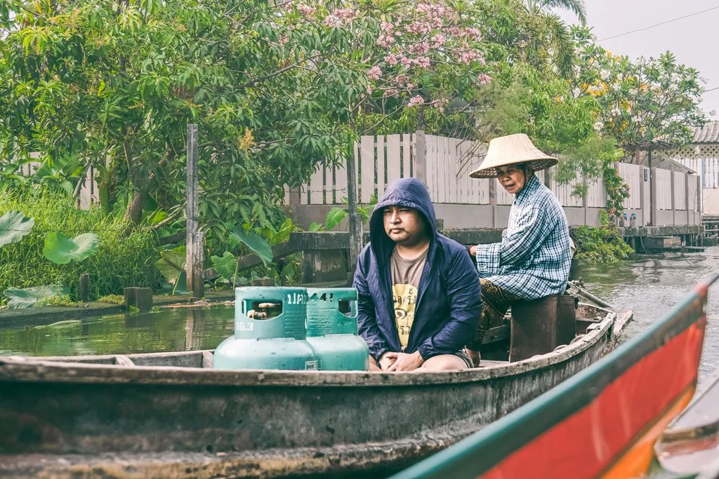 Two people in a small boat with a gas tank on a waterway, surrounded by lush green foliage and a wooden fence in the background.