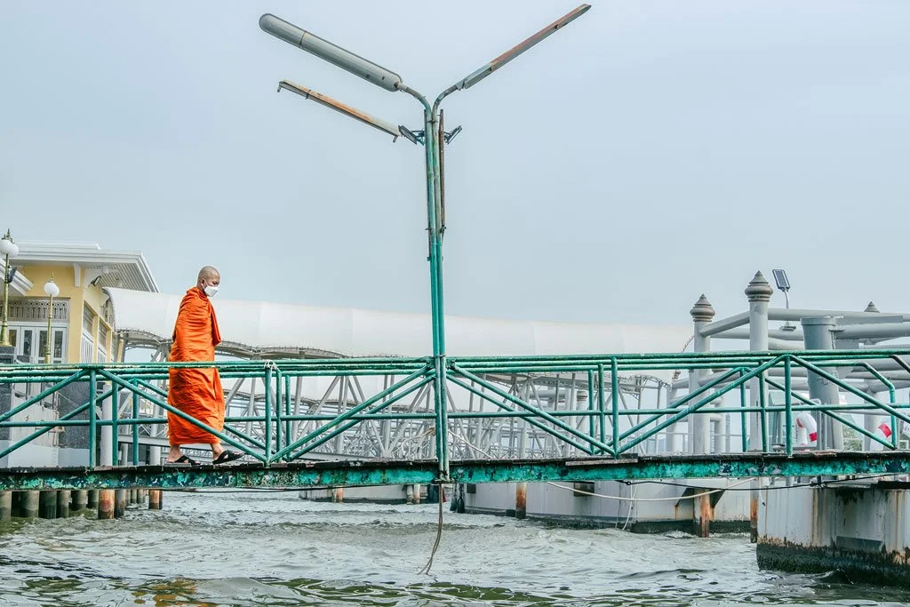 A monk walking on a floating bridge near a water plant, wearing a face mask and orange robes.